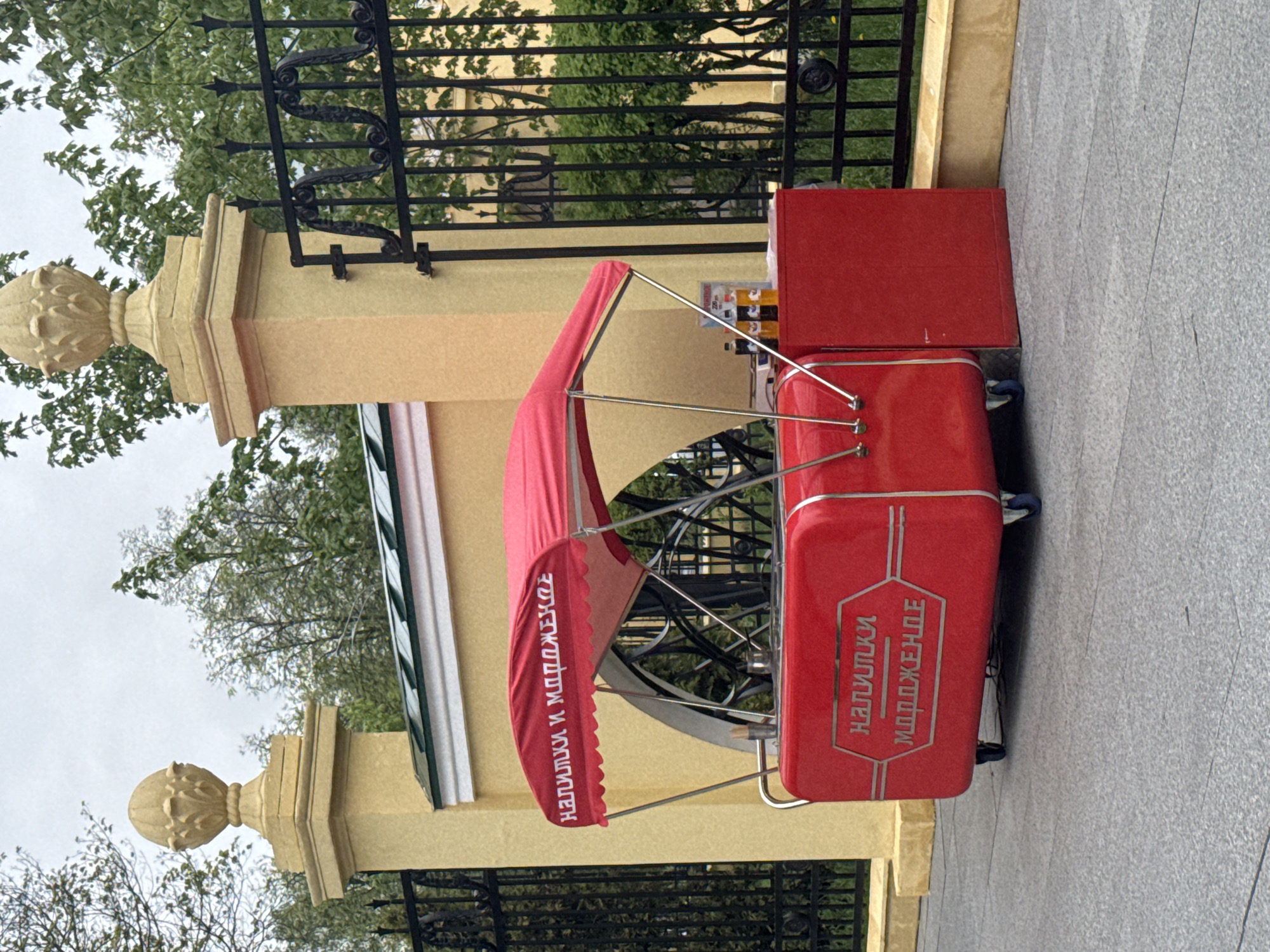 An eye-level, rotated, outdoor shot depicts a red vending cart next to a decorative yellow barrier fence on a gray-paved area in Nizhny Novgorod, Russia. The cart's red canopy reads "НАПИТКИ И МОРОЖЕНОЕ" in white text. The front of the cart features the same phrase in silver. Behind the cart, a black metal fence stands in front of green trees and a building. The barrier fence has yellow pillars topped with decorative spherical caps.