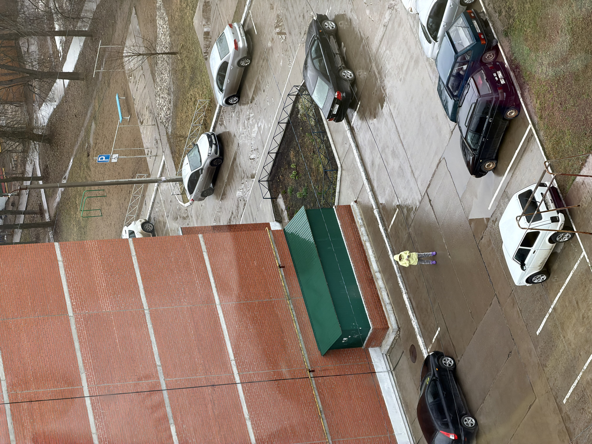 This high-angle, top-down image captures a residential courtyard and parking area in Chaikovsky, Russia, under overcast skies, likely during late winter or early spring. The ground is wet with puddles on the asphalt and concrete pavement, which is marked with white parking lines. Patches of dry, sparse grass and bare trees are visible, along with some remnants of melting snow or ice.
Multiple cars are parked throughout the scene. On the right, a white car (likely an older model Lada) and a dark blue/black sedan are parked parallel to a grassy strip. Further left, a silver sedan and another silver car are visible. A black sedan is parked closer to the bottom right, and a white hatchback is partially visible on the far left.
A single person is visible in the middle-right of the frame, standing on the wet pavement. They are wearing a light yellow/green hooded jacket, gray pants, and purple boots. Their activity is limited to standing.
Notable objects include a blue square parking sign with a white 'P' symbol, other white signs, and various metal poles and structures, some of which appear to be playground equipment (green bars, blue bench-like structure). A long, green, sloped-roof structure, possibly a shed or canopy, is situated next to a tall red brick wall on the left side of the frame. A yellow cable or hose runs along the ground near the brick wall.
Several car license plates are partially or fully visible. For instance, plates on some vehicles clearly show the Russian region code '152' (Nizhny Novgorod Oblast), such as "X328XM 152", "X855OX 152", and "Y196PO 152". The absence of strong shadows suggests an overcast day. The overall scene depicts a mundane urban environment.