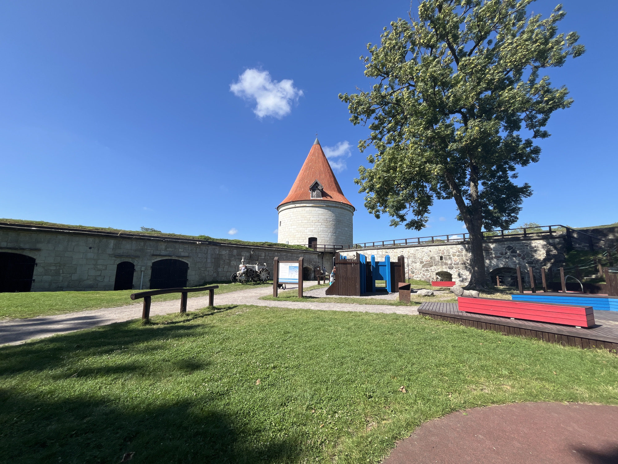This is an outdoor scene at the Kuressaare Episcopal Castle in Kuressaare, Estonia, on a clear, sunny day. In the foreground, a well-maintained green lawn is visible, with a curved reddish-brown pathway bordering its right side. To the left, a simple wooden fence runs along a path.
Dominating the mid-ground is a courtyard area with a playground structure featuring blue and brown elements. Adjacent to the playground are red and blue wooden benches. A large, leafy tree with green foliage stands prominently on the right side of the image, its branches extending towards the sky. Behind the tree, a section of the castle wall is visible, constructed from rough-hewn stones, with arched openings suggesting historical fortifications.
In the background, a circular stone tower with a distinctive orange-tiled conical roof rises against a bright blue sky. The tower has a small, dormer-like window on its upper section. The castle walls themselves are of stone construction, topped with a green, grassy embankment. A few white clouds dot the expansive blue sky. Historical artifacts, possibly cannons, are displayed near the castle walls. The overall impression is one of a well-preserved historical site with modern amenities, such as a playground, integrated into its grounds.