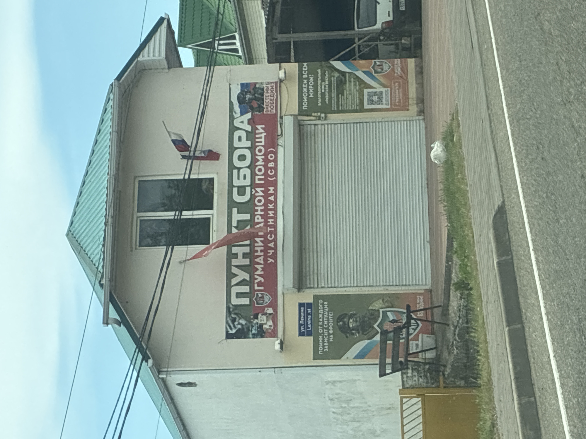 The image depicts a building in Arkhipo-Osipovka, Russia, with a prominent banner reading "ПУНКТ СБОРА ГУМАНИТАРНОЙ ПОМОЩИ УЧАСТНИКАМ (СВО)" which translates to "Collection Point for Humanitarian Aid for Participants (SVO)". The banner also features an image of a soldier. To the left of the banner is a sign indicating "ул. Ленина", meaning "Lenina St.", and below it a poster with the text "ПОМНИ, ОТ КАЖДОГО ЗАВИСИТ СИТУАЦИЯ НА ФРОНТЕ!" ("Remember, the situation on the front depends on everyone!") and an image of a soldier with Russian flags. To the right of the main entrance, which is a large, closed roll-up door, is another poster with the text "ПОМОЖЕМ ВСЕМ МИРОМ!" ("Let's help the whole world!"). A bench is positioned in front of the building. Two Russian flags are visible on the building. The scene appears to be a public or community space, possibly a storefront or administrative building, given the signage.