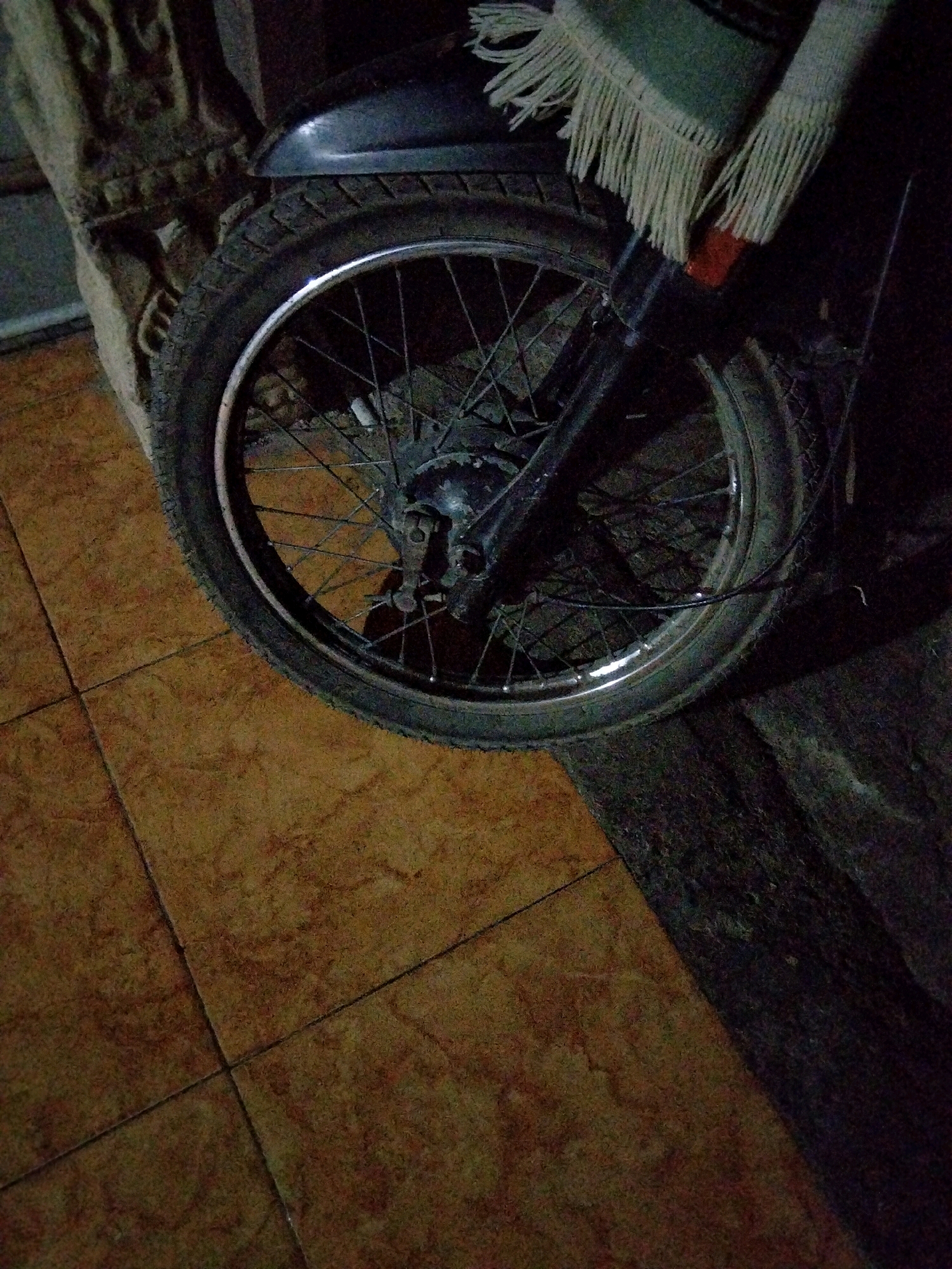 The image shows a close-up of a motorcycle wheel, its tire, and spokes. The motorcycle is parked on a tiled floor with a rough, dark surface to the right, possibly a road or pathway. In the background, a textured, light-colored object, possibly a pillar or wall with intricate carvings, is visible. A portion of the motorcycle's fender and a green textile with white fringes are also present in the upper part of the frame. The lighting suggests it is either dusk or an indoor setting with dim light. The image does not contain any people, and no specific location in Jenangan, Indonesia can be identified from the visual information.