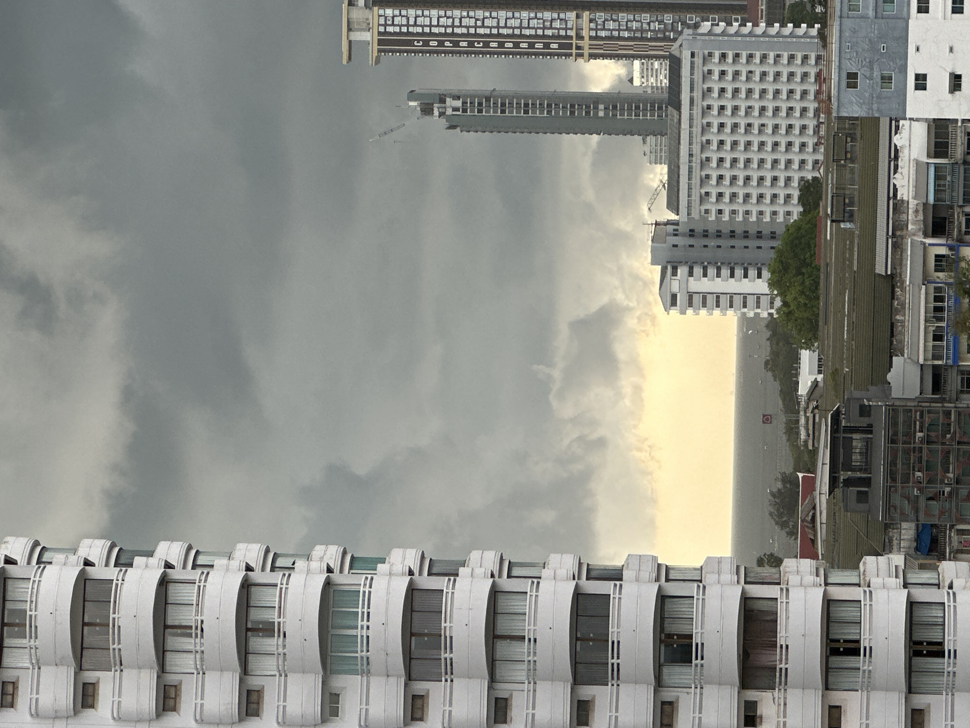 The image is an upside-down, wide-angle view of Pattaya City, Thailand. The foreground shows the white facade of a building with rounded balconies, which repeats across the bottom of the frame. Above this, the sky is predominantly grey with a patch of yellow, suggesting either a break in the storm clouds or a setting sun.
In the background, skyscrapers and other buildings stretch into the distance. The urban landscape includes a variety of architectural styles and building heights, indicative of a densely populated city center. The overall mood is muted, likely due to the overcast weather conditions.