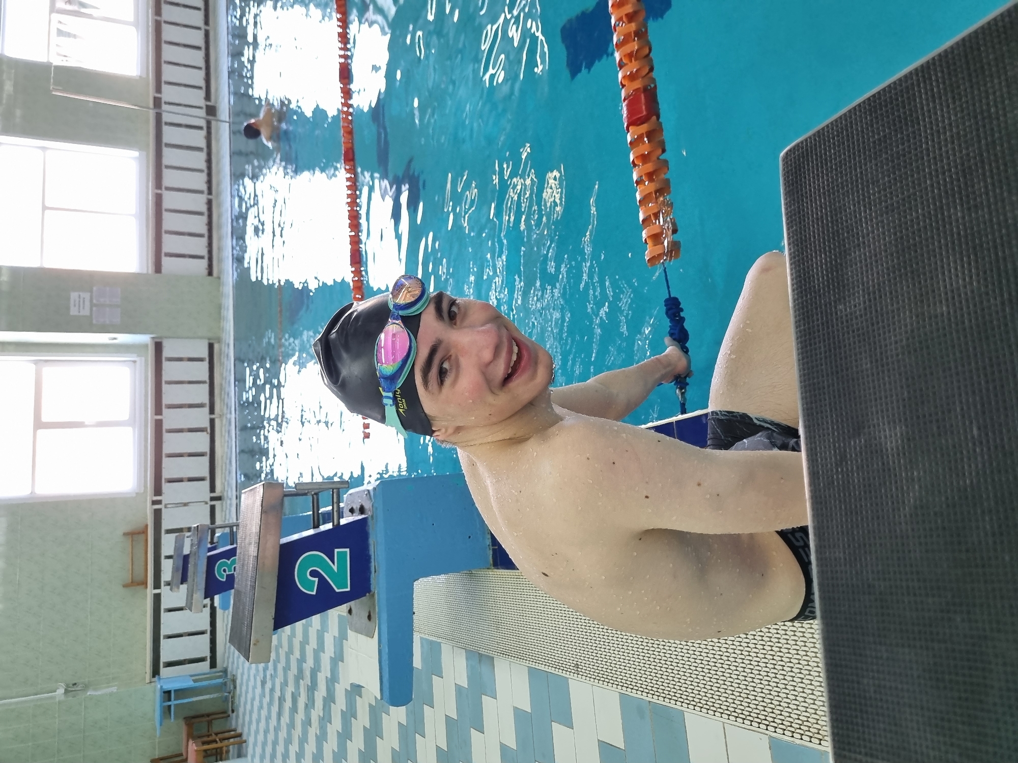 A young man wearing a black swim cap and goggles sits on the edge of a swimming pool, possibly in Markovskii, Russia. He is holding onto a lane divider with orange floats. He is smiling and looking at the camera. Another person is swimming in the background. The pool has lane markers and starting blocks. The pool area has tiled walls, windows, and tiled floors.