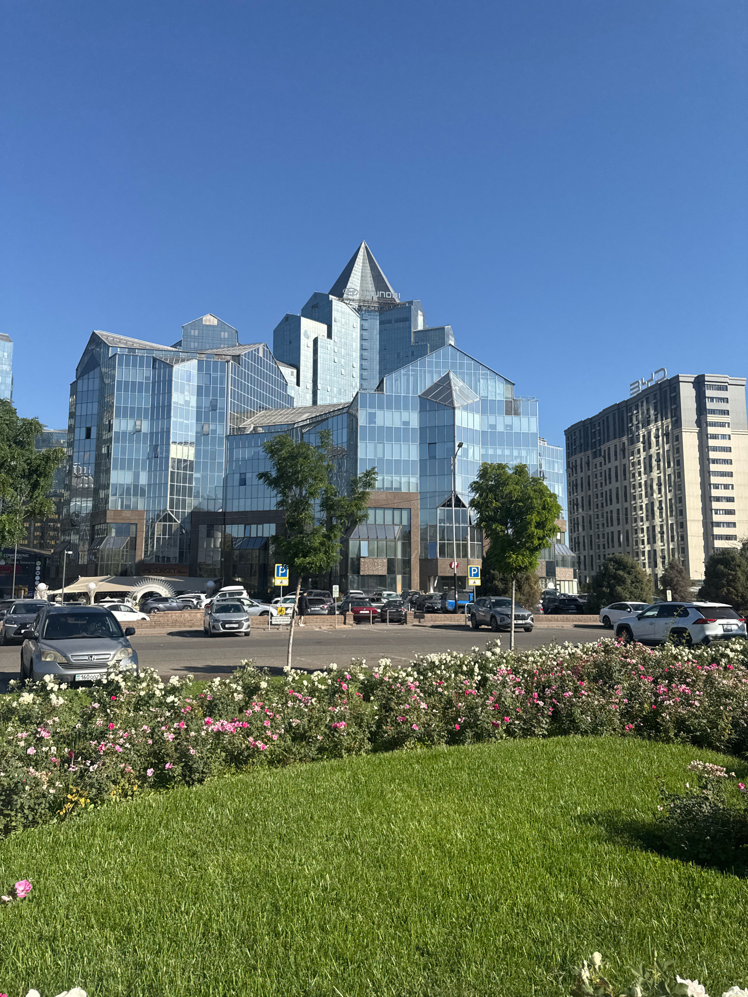 The image shows a modern cityscape with a prominent glass-fronted building complex in the foreground, likely a commercial or office space. This building features a distinctive faceted roofline and large windows reflecting the clear blue sky. To the right of this building is a taller, more rectangular building with a lighter facade and numerous windows. In front of these structures, a street is visible with several cars parked and in motion. A flowerbed filled with pink and white roses lines the foreground, adjacent to a well-maintained green lawn. The overall scene suggests a sunny day in a well-developed urban area. This building complex is identified as the "Nurly Tau" business center in Almaty, Kazakhstan, with its unique architectural design making it a recognizable landmark in the city.