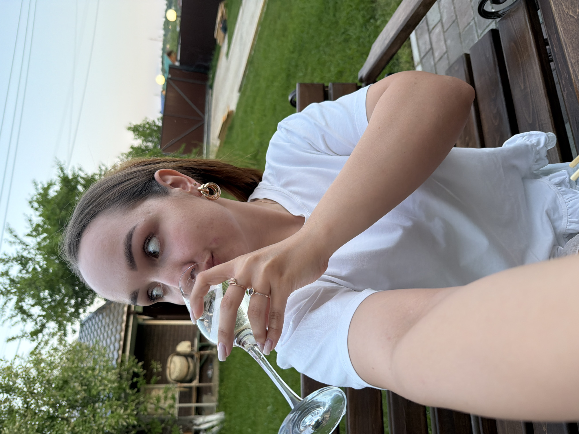 A young woman is holding a glass of champagne. She is sitting on a wooden bench in a backyard with a lawn. The location is the city of Chaikovsky, Russia. In the background, there's a fence and some greenery. The woman is wearing a white shirt and gold earrings, and she has two rings on her fingers. She is looking into the camera while holding the glass.