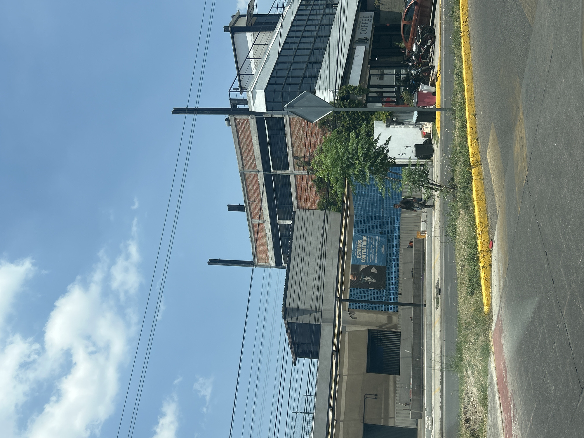 The image depicts a street scene in San Bernardino Tlaxcalancingo, Mexico, featuring a building under construction with exposed brick and concrete framework. A multi-story building with a "COFFEE" sign is visible nearby. Along the sidewalk, there's a blue advertisement banner, a small tree, and a person walking. The street has a yellow curb and is lined with vegetation. Overhead, power lines crisscross the sky, which is partially cloudy.