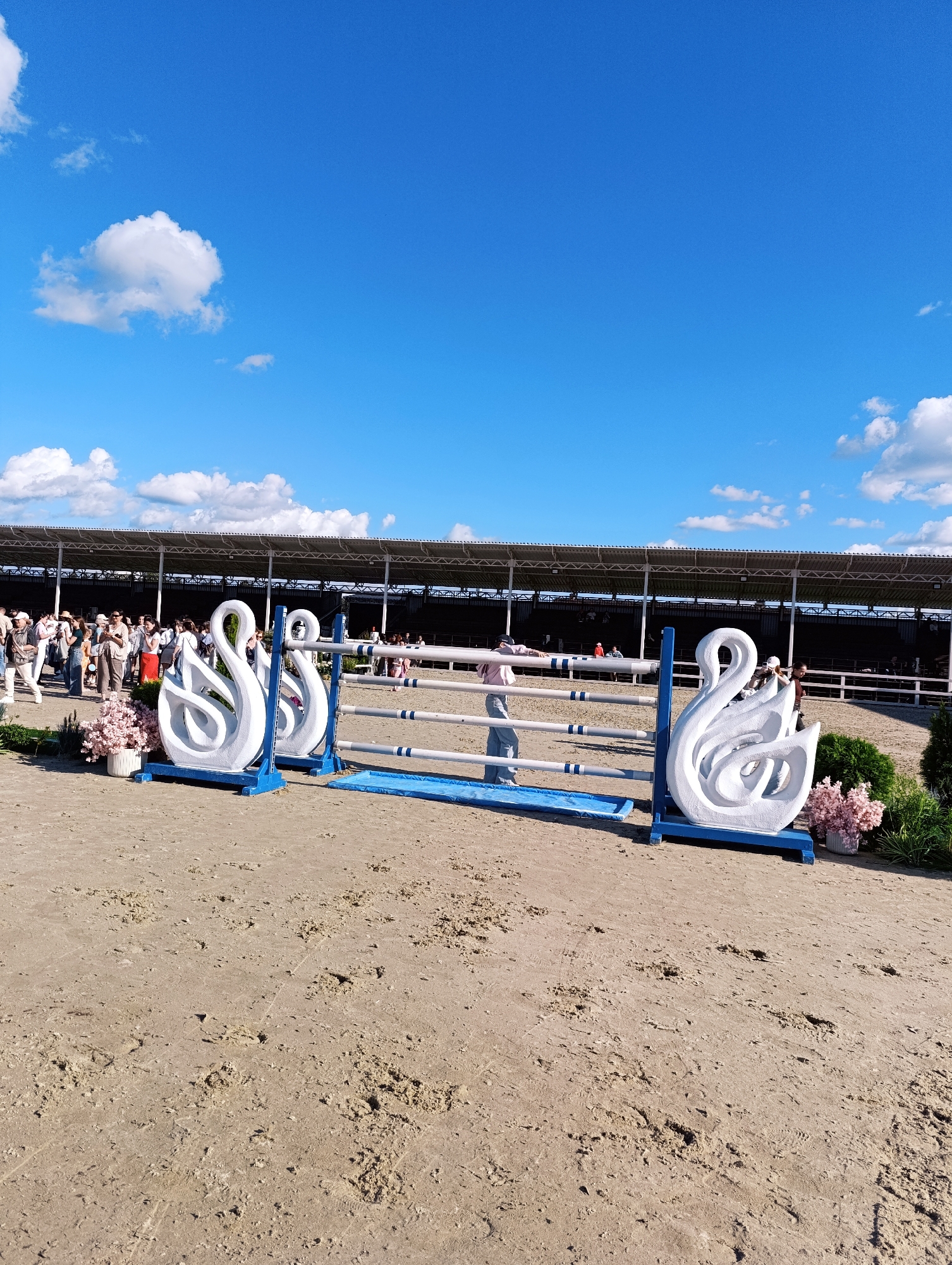 This is a medium shot of a horse jumping arena on a sunny day in Ivanovskoe, Russia. The sky is bright blue with scattered white clouds. In the foreground, a sandy arena floor is visible with some footprints and divots. A horse jumping obstacle is set up, featuring two white swan-shaped supports. The obstacle itself is made of white poles with blue bands. To the left of the obstacle, there are pink floral decorations. In the background, a grandstand with a roof is visible, and a crowd of spectators is gathered, some standing and others seated. Several people are milling about near the arena. A person in a pink top and jeans is leaning on one of the obstacle poles. The overall scene suggests an outdoor equestrian event.