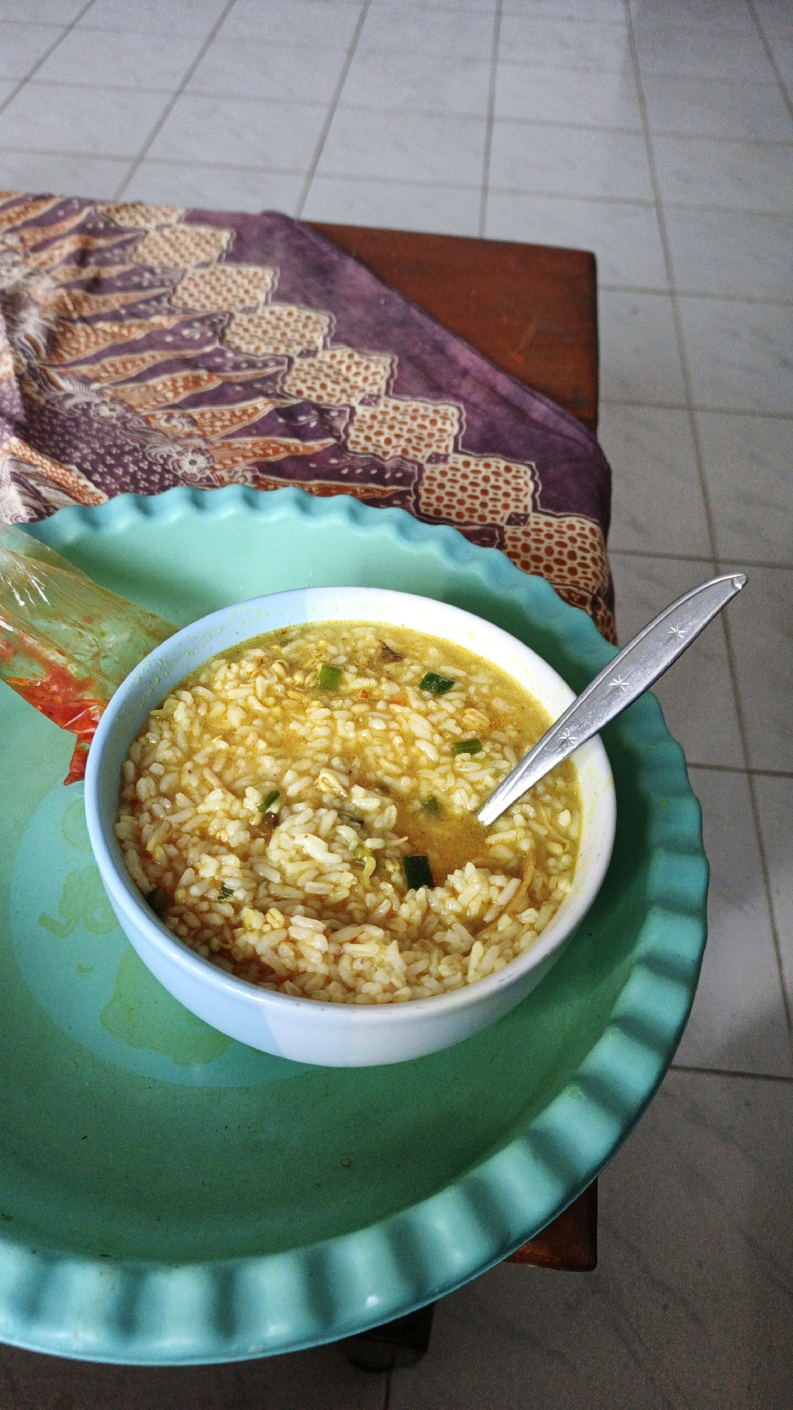 A close-up, high-angle shot captures a bowl of congee, a rice porridge, served on a teal-colored tray. The congee, which appears to be a savory dish with visible rice grains and flecks of green onion and other ingredients, is topped with a glistening broth. A silver spoon rests in the bowl. To the left of the bowl, a clear plastic packet contains a red sauce, likely chili. The tray and bowl are placed on a wooden surface, partially covered by a piece of fabric with a purple and orange patterned design. In the background, a tiled floor is visible. This meal is likely a typical breakfast or light meal, commonly found in the rural settings of Campurdarat, Indonesia, where such dishes are a staple.
