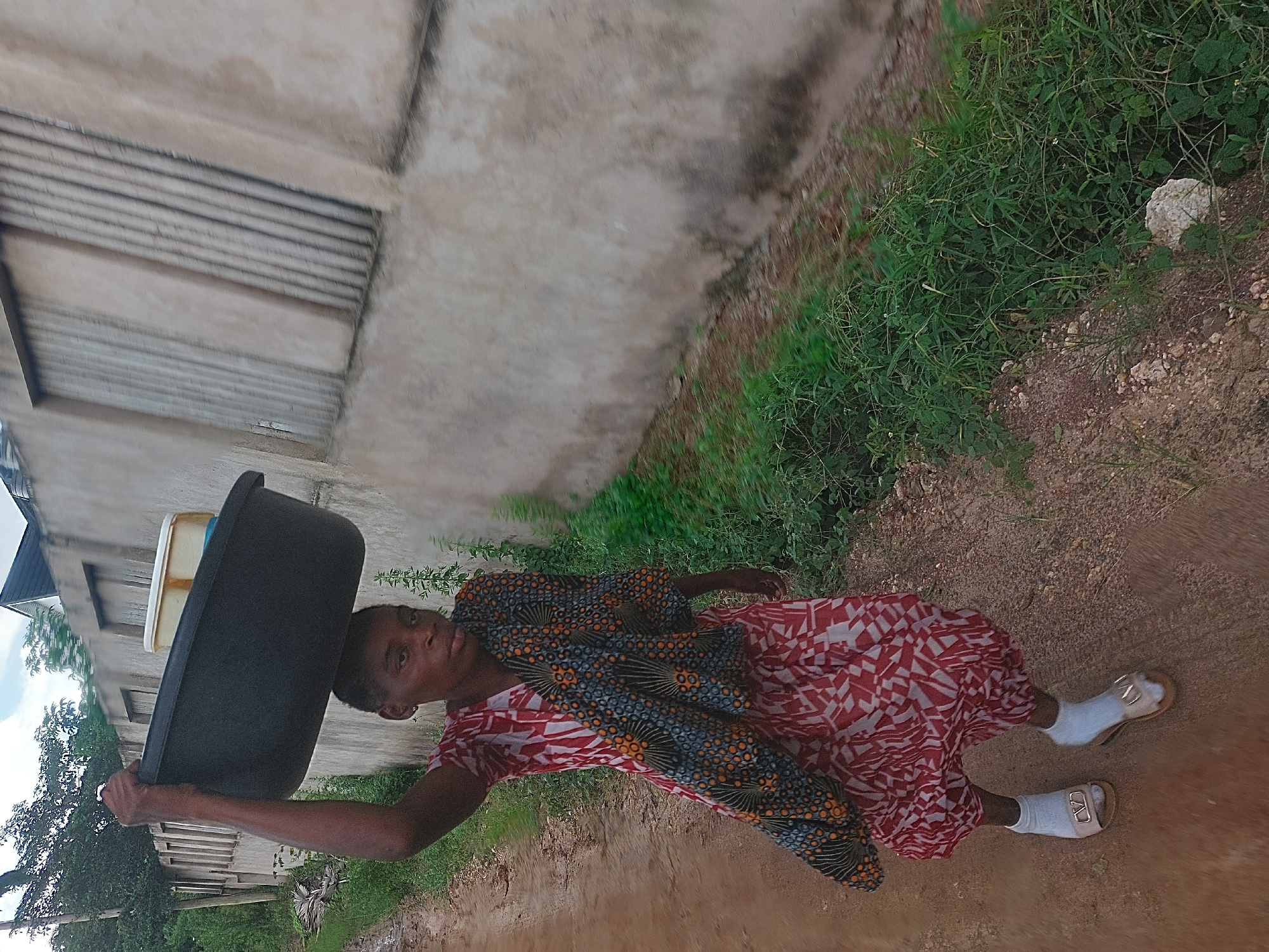 A full-body shot captures a woman walking on a dirt path in what appears to be a residential or semi-urban area of Dar es-Salaam, Tanzania. She is carrying a large, dark grey or black plastic basin on her head, steadying it with her right hand. Inside the black basin is a smaller, cream-colored rectangular container.
The woman has short, dark hair and is looking directly at the camera with a neutral expression. She is dressed in a knee-length dress featuring a vibrant red and white geometric pattern. A piece of fabric, dark blue with orange and yellow circular patterns, is draped over her shoulders and upper torso. On her feet, she wears white socks paired with light-colored, open-toed sandals that have a distinct gold-toned buckle on each.
The background includes a light-colored concrete or block building with horizontal grey lines or grooves visible on its facade. To the right of the dirt path, there is an abundance of lush green vegetation and reddish-brown soil, characteristic of unpaved areas. A small, irregularly shaped white rock is visible nestled within the green foliage. The lighting suggests it is daytime, possibly under an overcast sky or during the late afternoon. There is no visible text in the image.