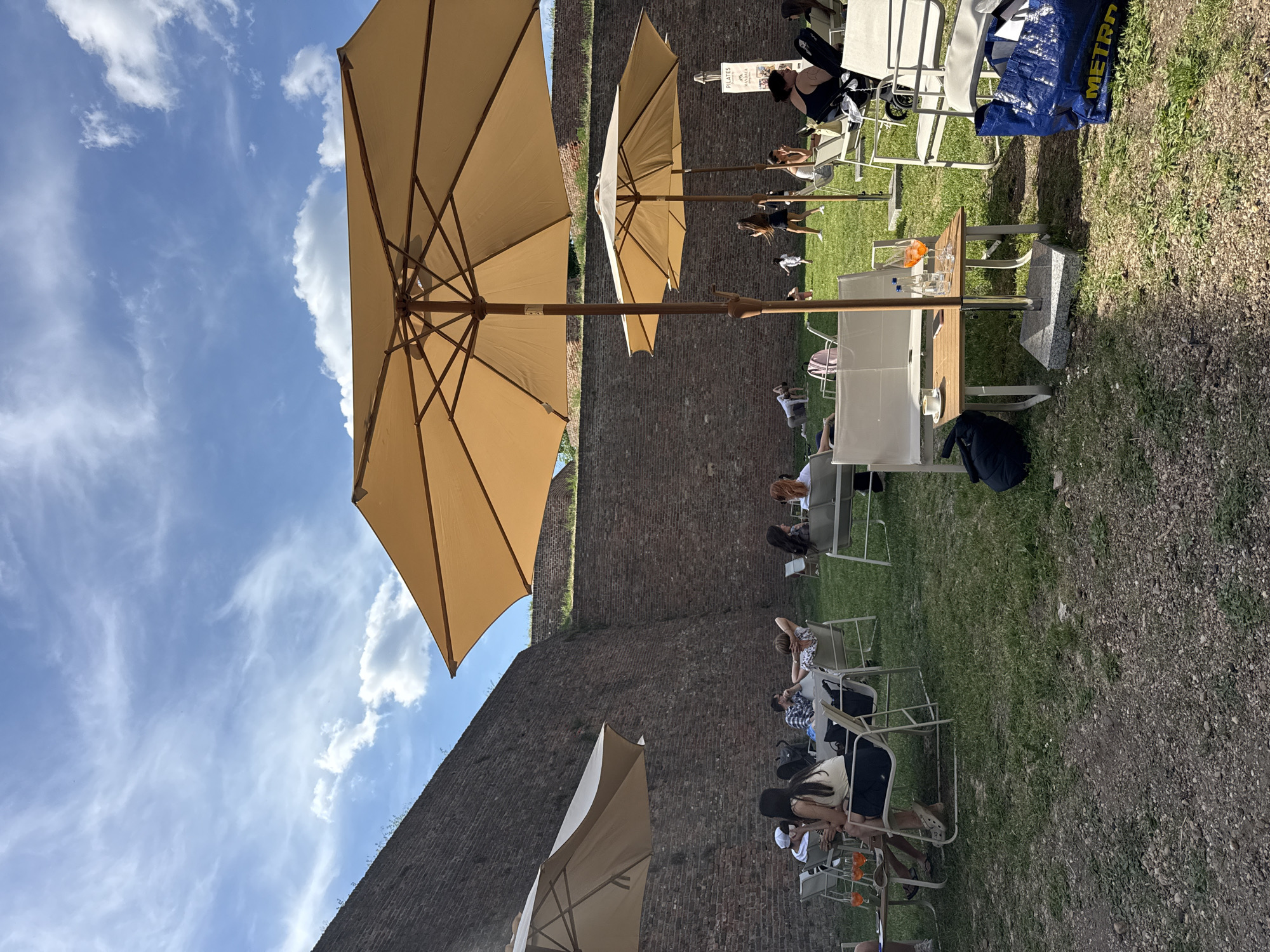 The scene is an outdoor seating area in Belgrade, Serbia, adjacent to an old brick wall. Several tan-colored umbrellas provide shade for patrons sitting at tables. People are seated at the tables, some engaging in conversation, while others are relaxing. A few chairs are unoccupied. The ground is covered with grass and gravel. The sky is blue with scattered white clouds.