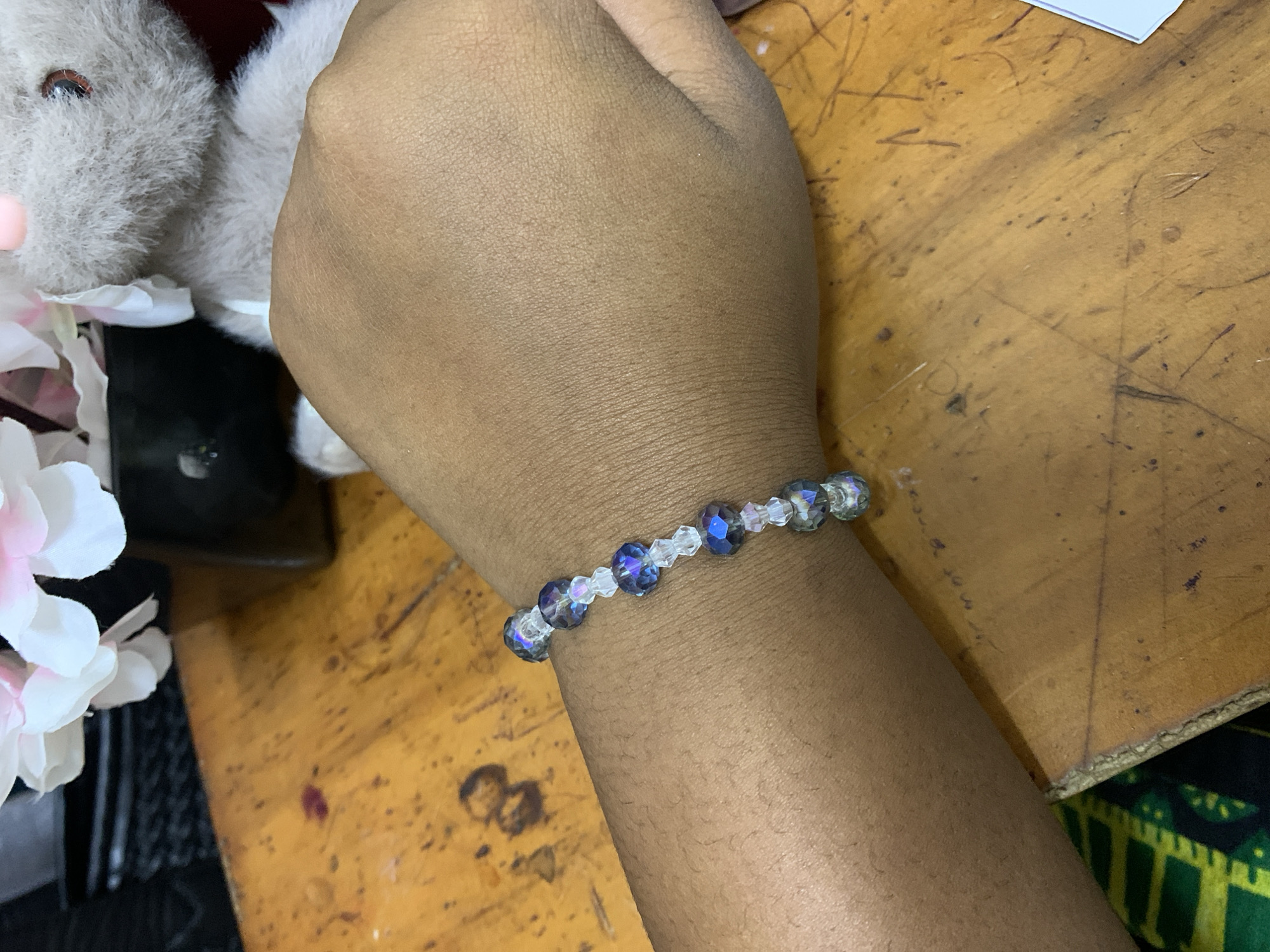 A tan-skinned arm wearing a blue and clear beaded bracelet is centered in the frame, positioned against a worn, light brown wooden surface. The arm leads to a hand that is partially obscured by a gray stuffed animal and some white artificial flowers. A black object is also visible behind the flowers. The wooden surface shows signs of wear, including scratches and marks. The bottom right corner reveals a black surface with a green and yellow geometric pattern. Without further information from the image, identifying the exact location within Dar es Salaam, Tanzania, is not possible.