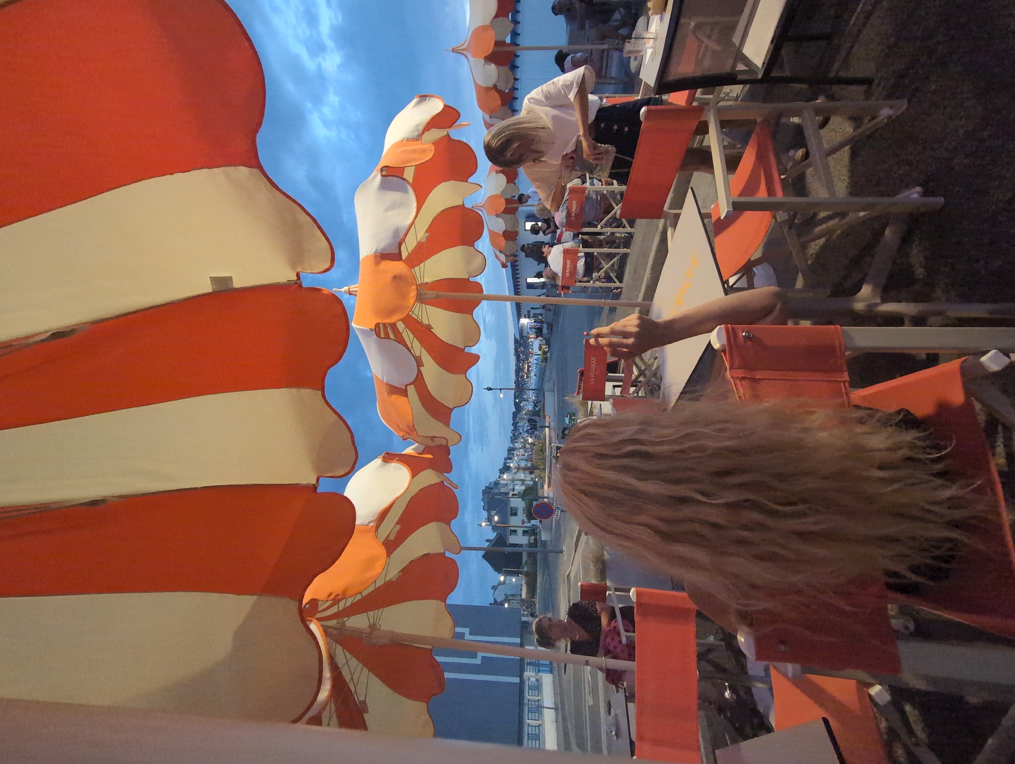 The image captures an outdoor dining scene under striped orange and white umbrellas, likely in Mers-les-Bains, France, given the architectural style of the buildings in the background and the presence of beach umbrellas. In the foreground, a person with long blonde, curly hair is seated facing away from the camera. Adjacent to them, another person with blonde hair and a white shirt is attending to a table, appearing to put something into a bag. Several other patrons are visible seated at tables in the middle ground, under similarly colored umbrellas. The setting appears to be a promenade or street next to a beach or waterfront, with buildings and streetlights lining the area. The sky is overcast and a dusky blue, suggesting it is either early morning or late evening. The overall impression is of a relaxed, perhaps casual dining environment in a coastal town.