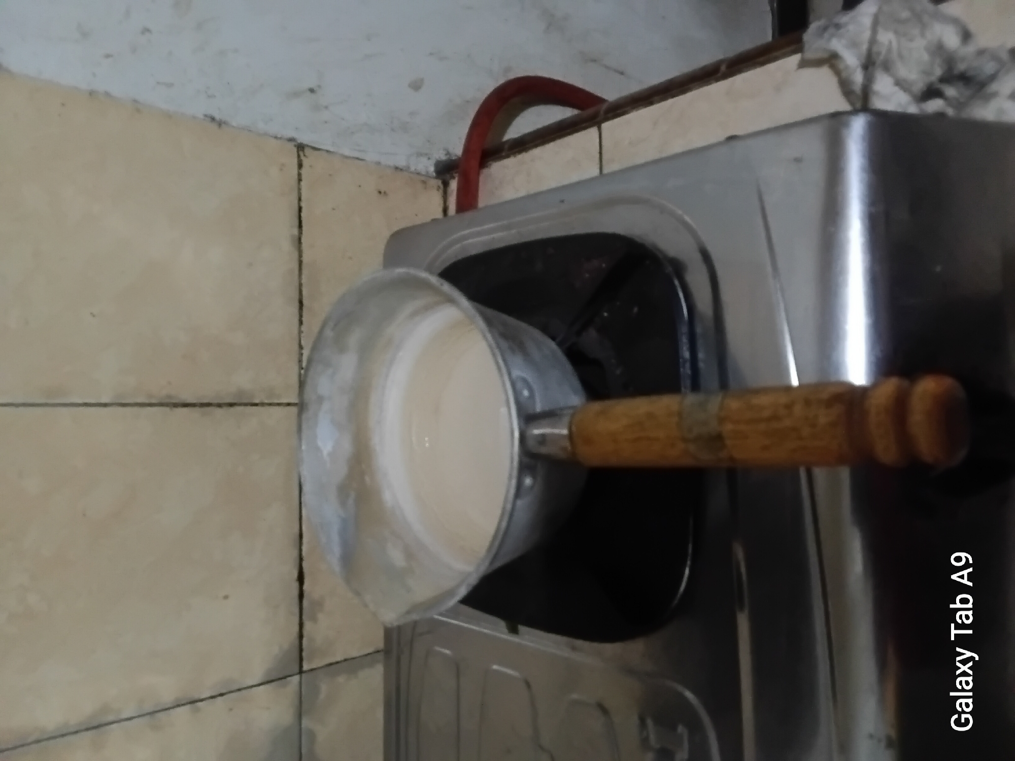 A pot filled with a light-colored liquid sits on a gas stove. The pot has a wooden handle. The stove is a metallic, two-burner model with a black grate. Behind the stove, the wall is tiled with light brown square tiles. A red gas pipe is visible to the right of the stove, leading up towards the wall. The floor appears to be made of light-colored tiles. A crumpled white cloth is on the floor to the far right. The scene is indoors, likely in a kitchen. The image is watermarked with "Galaxy Tab A9" in the lower left corner.