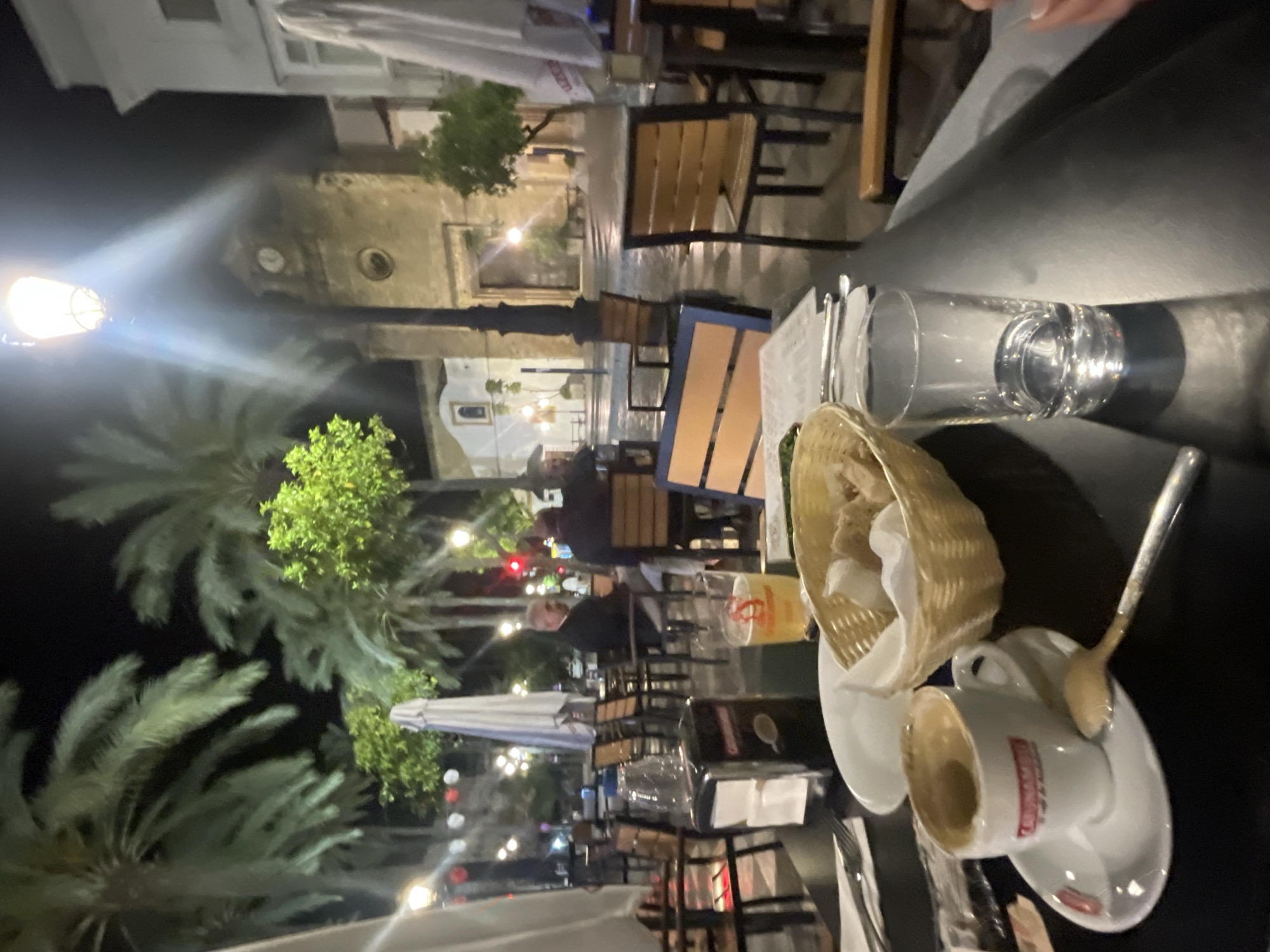 The image depicts an outdoor dining scene in Jerez De La Frontera, Spain, at night. The view is from a table, with a half-empty coffee cup on a saucer, a bread basket, a spoon, and a glass of water in the foreground. Tables and chairs are visible in the middle ground, occupied by people dining. In the background are palm trees, streetlights, and architectural structures, including a building with a clock. Umbrellas are present, suggesting an outdoor seating area. The scene is illuminated by artificial lights.