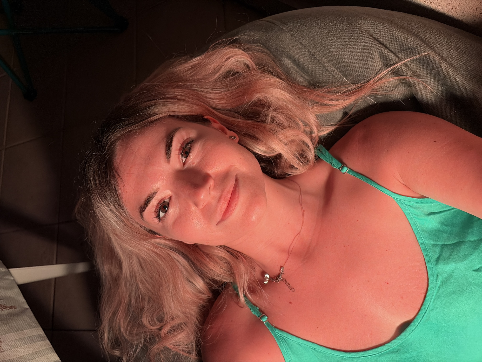 A close-up, high-angle selfie features a young woman with long, wavy blonde hair tinged with pink lying down. She is wearing a bright green tank top with thin straps. A delicate silver necklace with a small pendant rests on her collarbone. Her right eye is visible, looking towards the camera. The lighting casts a strong orange hue across her skin. In the background, a dark, textured surface, possibly a couch or chair, is visible, along with hints of a striped fabric and what appears to be furniture legs. The scene is dimly lit, with shadows dominating the upper portion of the image. No specific location in Budva, Montenegro is identifiable.