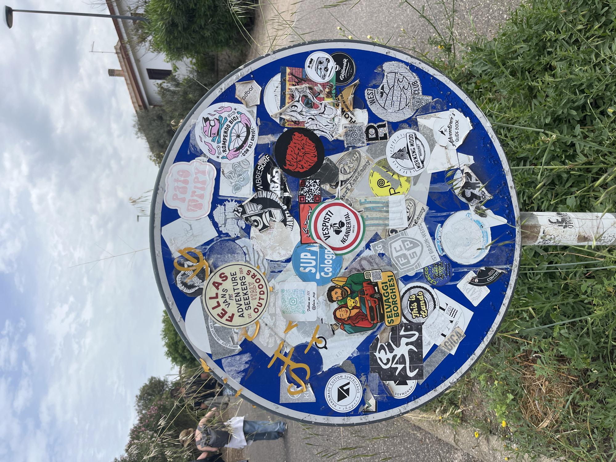 The image shows a blue circular signpost, densely covered with a variety of stickers. The sign is located in what appears to be Santeru/San Vero Milis, Italy, as indicated by text on one of the stickers.
The stickers display various designs, logos, and text, including references to adventure seekers, surfing, travel, and local Italian culture.
The background includes foliage, a paved road, and a glimpse of a building. Some people are visible near the bottom left of the image, suggesting they are walking on the road next to the signpost.