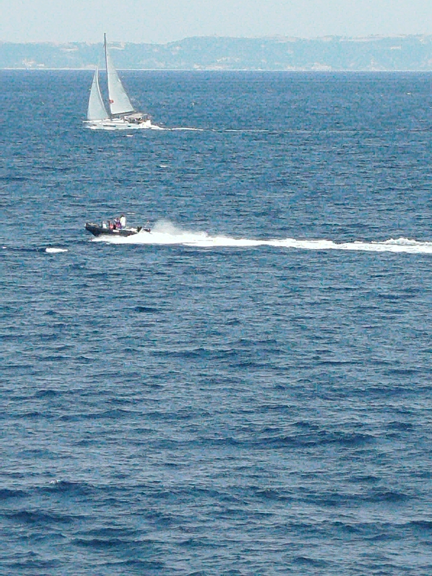 In a wide shot of the sea, a white sailboat with two sails is visible in the upper left quadrant, moving from left to right. In the lower middle of the image, a smaller black motorboat with several people onboard is traveling from left to right, creating a white wake. The water is a deep blue with visible ripples. In the background, a hazy, light-colored landmass with hills can be seen on the horizon. The sky is a pale, overcast blue. The scene appears to be on the open sea.