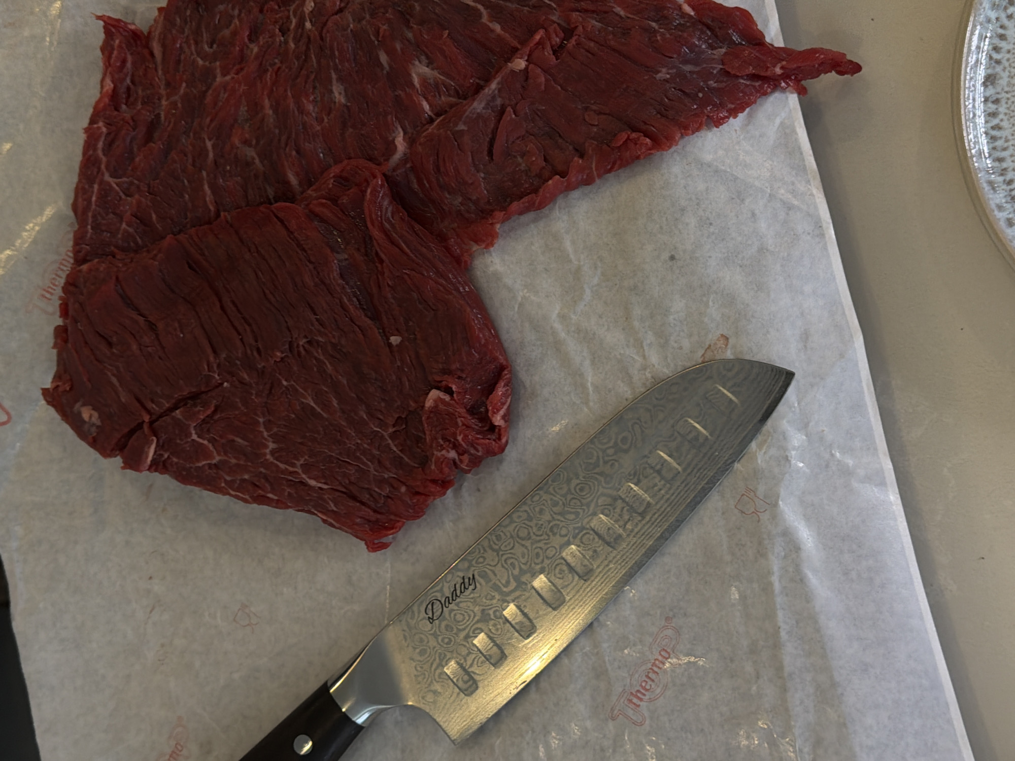 The image displays several pieces of raw, thinly sliced red meat, likely beef flank or skirt steak, laid out on a sheet of white parchment paper. The meat is a deep red color, showing muscle fibers and irregular shapes, indicative of freshness and preparation. Next to the meat lies a gleaming chef's knife, possibly a Santoku style, featuring a distinctive patterned blade, characteristic of Damascus steel, and a dark handle. The word "Daddy" is visibly engraved in a cursive font on the knife's blade.
The parchment paper has faint red printed details, including a partial word "Thermo" and a small, stylized logo that appears to be a cooking symbol. A portion of a light-colored, possibly ceramic, plate with a textured or speckled pattern is visible in the upper right corner of the frame. The scene depicts an indoor setting, most likely a kitchen or food preparation area, where the raw meat is being prepared for cooking or further processing. No specific time of day, weather conditions, or emotions are discernible. The image provides no visual cues to suggest the specified location context of London, United Kingdom.