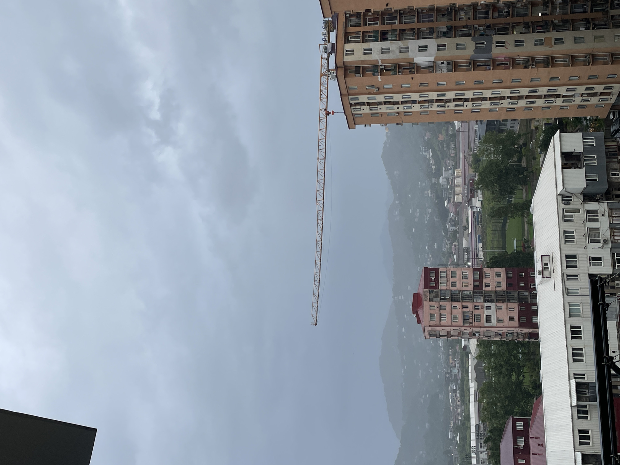 This media file shows a view of a city skyline with multiple buildings and a construction crane. The buildings are of varying heights and architectural styles, with some appearing to be older apartment blocks and others more modern structures. One prominent building is a tall, beige apartment building with many windows. To its right, a bright yellow construction crane extends diagonally across the frame, its long arm reaching towards the sky. Below the crane, a smaller, pink and red apartment building is visible, with a distinctive red roof. On the right side of the image, a modern white building with numerous windows is prominent. In the background, a dense urban landscape stretches out, with more buildings and lush green trees visible, nestled against a backdrop of misty, rolling hills. The sky is overcast and appears to be a dull gray, suggesting a cloudy or possibly rainy day. The image is oriented vertically, with the sky taking up the top half of the frame. There is no visible text in the image. The overall scene evokes a sense of urban development and a somewhat muted atmosphere due to the weather. The location appears to be Batumi, Georgia, given the context provided.