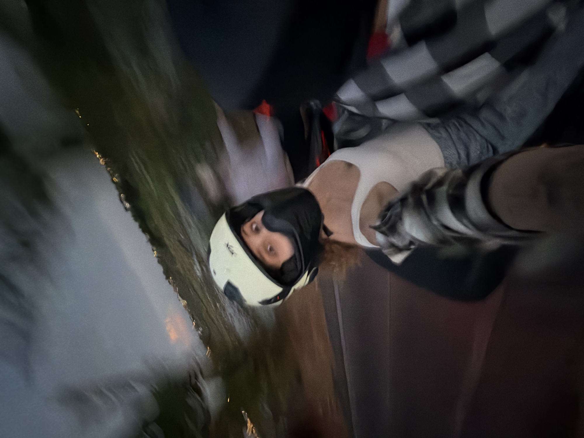 A person wearing a white and black helmet is visible in the image. The background shows what appears to be a city at night. Based on the information provided, the location is North Kuta, Indonesia. The person is wearing a white tank top and a black and white checkered jacket. Due to the blurred nature of the image, specific actions or interactions cannot be determined.