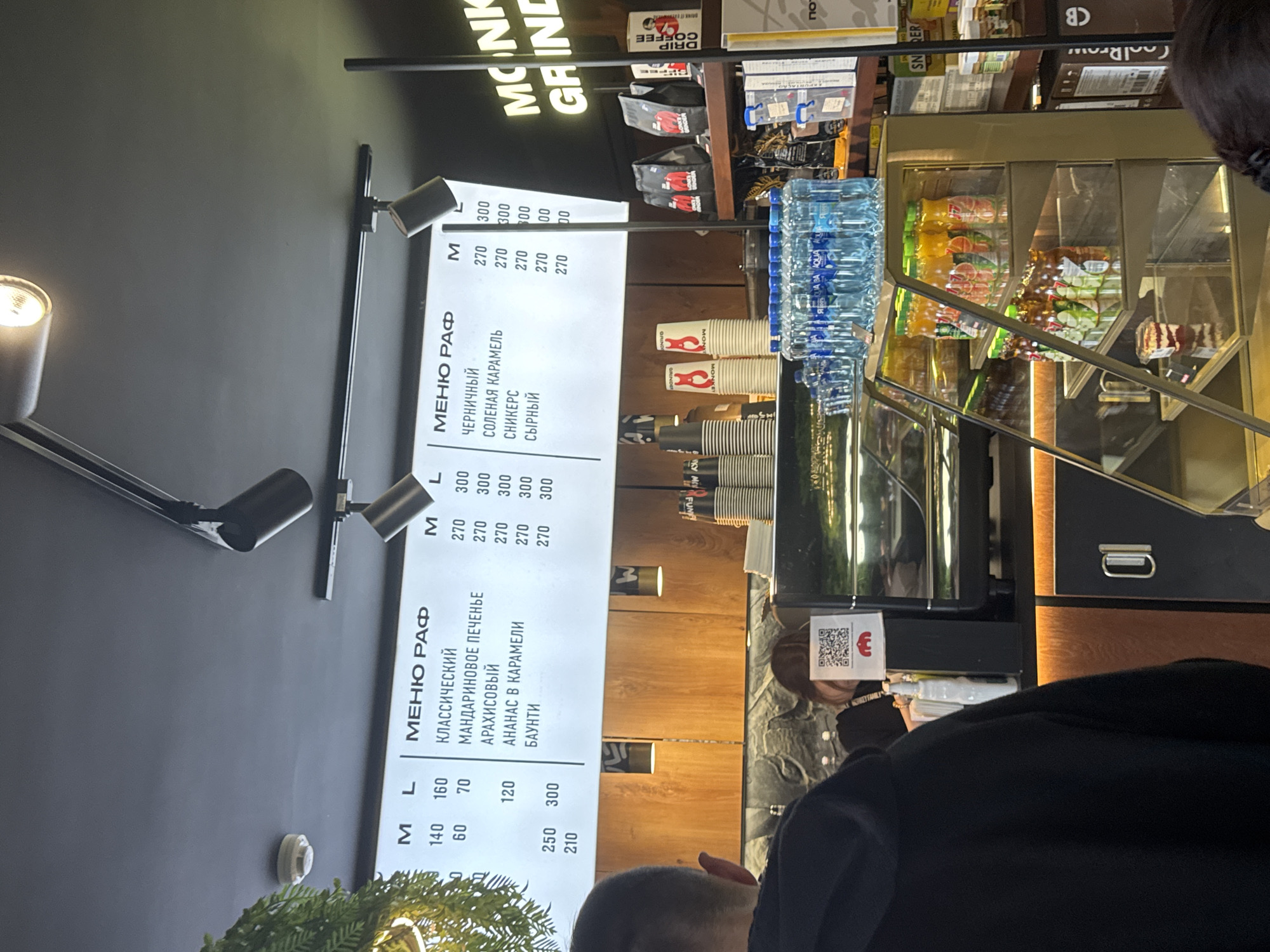 The image shows a coffee shop interior, likely in Perm, Russia, given the Cyrillic script on the menu board. The menu lists "RAF" (a coffee drink) with different flavors and prices for medium (M) and large (L) sizes. Bottles of water are stacked above a service area with coffee cups. A customer in a dark jacket is partially visible, standing near a counter. The brand "MONKI GRIND" is visible at the top, and there are various packaged food and beverage items on shelves.