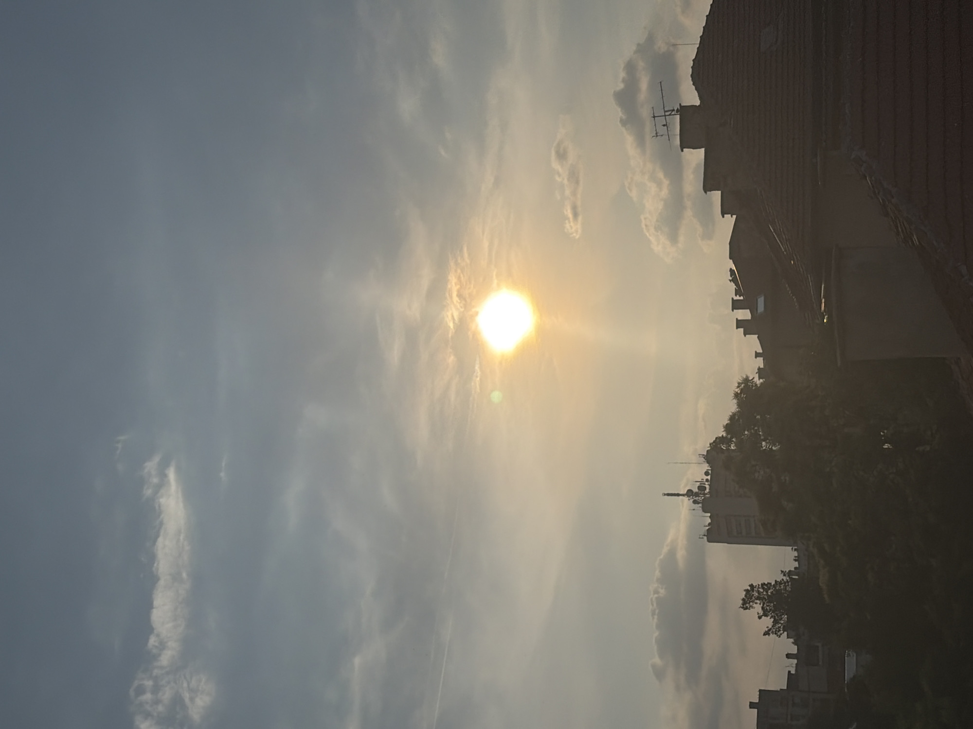 The image displays an urban landscape under a partially cloudy, pale blue-grey sky, illuminated by a low-lying sun, indicating either sunrise or sunset. The sun, a brilliant white-yellow orb, is positioned slightly to the left of the vertical center and about two-fifths down from the top, exhibiting strong lens flare and visible light rays. Scattered light grey and white clouds are distributed across the sky, with a distinct, thin horizontal contrail visible in the upper-left quadrant.
The lower third of the scene is dominated by the dark silhouettes of buildings and dense tree canopies. On the right, a cluster of residential rooftops with terracotta tiles and multiple chimneys is visible. A prominent television antenna, silhouetted against the sky on one of the closer rooftops, has a small, dark bird perched on its structure. To the left, a dense mass of dark-foliaged trees occupies the mid-ground. Behind these trees, in the mid-left, stands a taller, light-colored building featuring numerous communication antennas and equipment on its roof. Other indistinct building structures are partially visible in the far-left background. No human figures or discernible activities are present in the scene. The location is in the city of Unknown, Unknown.