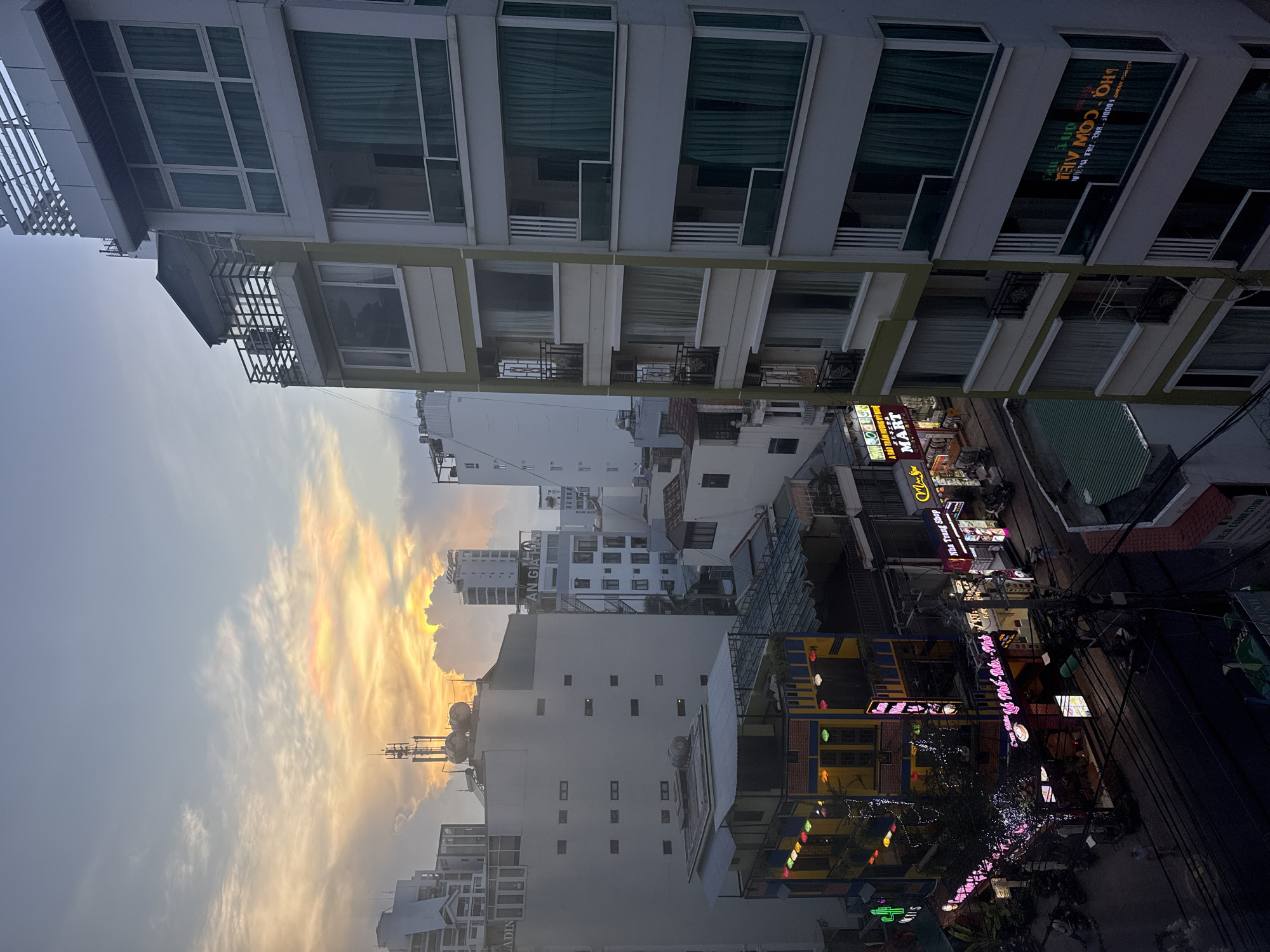 The image captures a cityscape, possibly of Nha Trang Ward in Vietnam, during what appears to be a colorful sunset or sunrise. The sky is a blend of soft hues, with orange and yellow gradients visible through the clouds.
In the foreground, there are various buildings ranging in height, typical of urban settings. The buildings closer to the viewer are multi-story structures with numerous windows and balconies. Signage in Vietnamese is visible on some of the buildings. The architecture is a mix of styles, some appearing modern while others seem older and more traditional.
At street level, there's evidence of commercial activity, with neon signs and various storefronts. These signs suggest a mix of businesses, including restaurants or eateries, shops, and possibly entertainment venues. Power lines stretch across the scene, adding to the urban feel. The area appears lively, with potential activity occurring in the businesses.