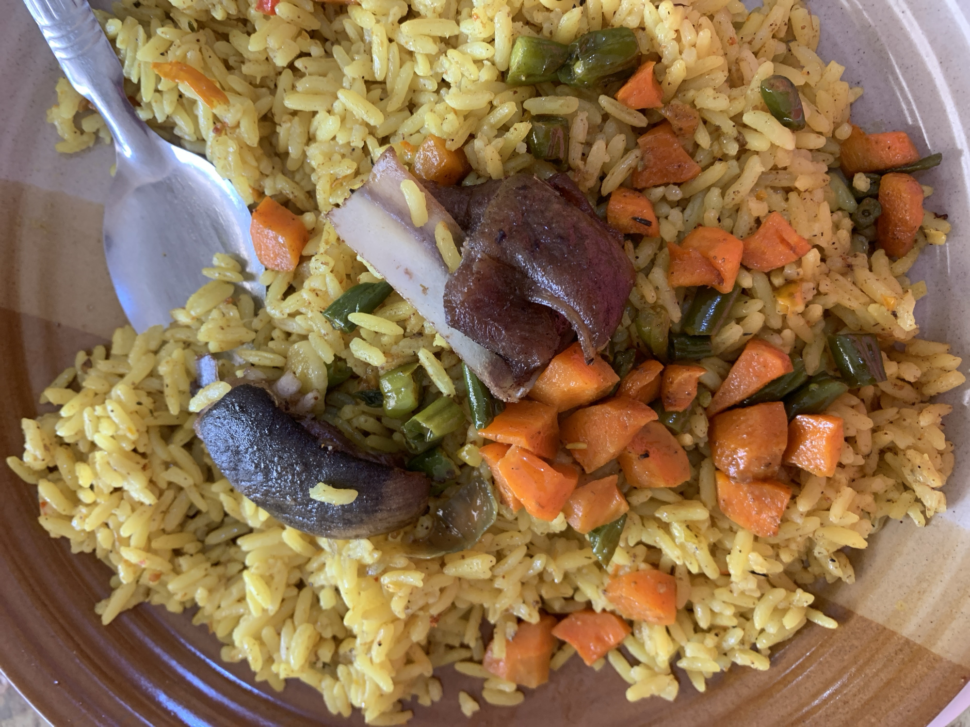 The image shows a plate of yellow rice with diced carrots, green beans, and two pieces of meat. A metal spoon rests on top of the rice. The plate has a beige and brown rim. The image was taken in Kabong, Nigeria.
