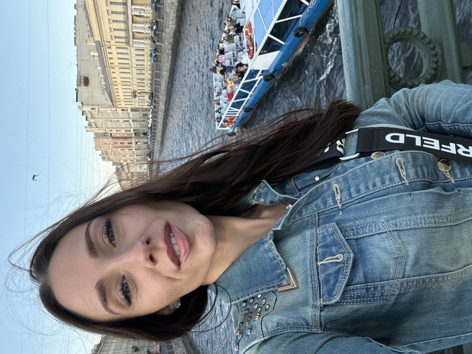 A woman takes a selfie on a bridge over a canal in Saint Petersburg, Russia. She has long dark hair, blue eyes, and is wearing a denim jacket with silver stud accents on the shoulders. Behind her, a blue and white tour boat filled with passengers navigates the canal, passing by ornate classical buildings that line the waterfront. The sky is clear and blue, with a few birds flying in the distance. The scene suggests a pleasant day for sightseeing in the city.