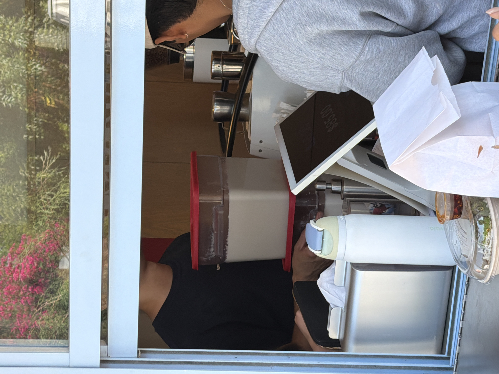 This image captures a vibrant scene at what appears to be a service window or stand, likely a coffee or food establishment, viewed from the outside looking in. The original photo is rotated 90 degrees counter-clockwise.
Inside the window, two individuals are present. One person, wearing a grey hooded sweatshirt, is visible from the chest up, leaning over equipment, suggesting they are a barista or server. They have short dark hair and a small earring. Partially obscured by the counter, another person wearing a black t-shirt is visible.
The counter space is filled with various items. Prominently displayed is a digital tablet screen showing "$65.00", indicating a transaction or pricing. Next to it are two white paper bags, possibly for food items or beverages. A large, clear container with a red lid holds a light-colored liquid, possibly milk or a beverage base, positioned centrally on the counter. To its right, a white water bottle with a blue and light-yellow cap is visible, with the word "solo" partially legible on its side. Below this, metallic elements of what looks like coffee or beverage preparation equipment are visible, alongside small clear jars containing spices or toppings.
Through the window on the left, an outdoor scene unfolds, featuring lush green foliage and a bush adorned with bright pink/purple flowers, suggesting a pleasant, natural environment. The presence of natural light indicates it is daytime. The overall setting implies a casual, service-oriented interaction, possibly in an urban or semi-urban outdoor area like Puebla, Mexico.