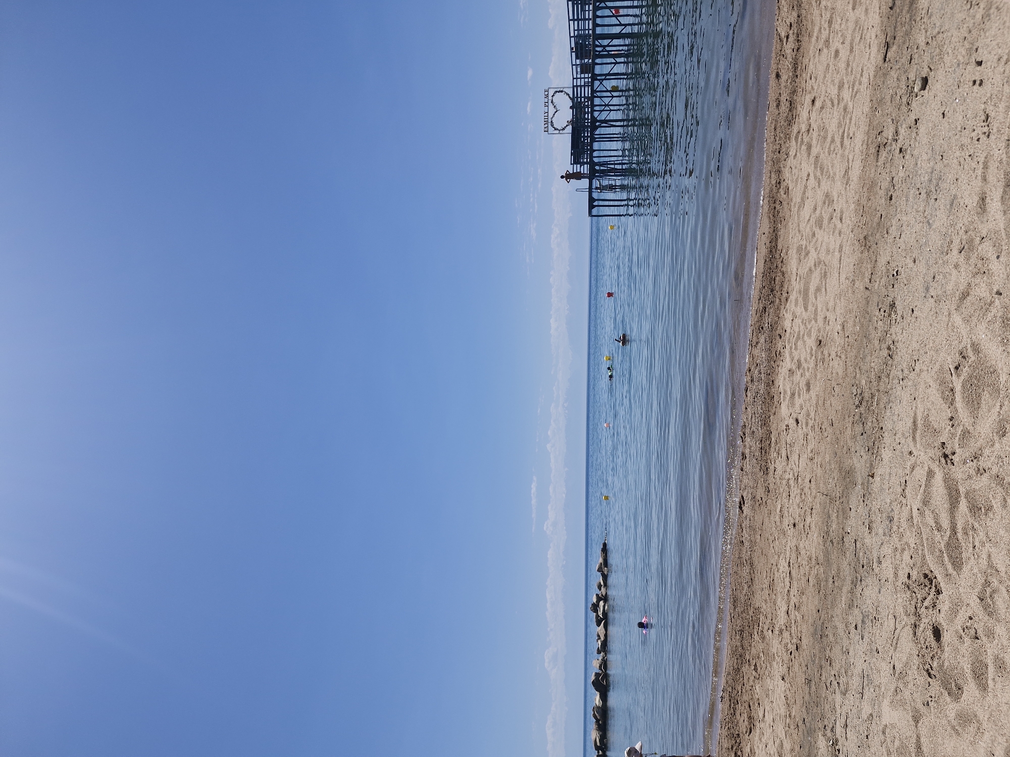 This image depicts a clear, sunny day at a beach on Issyk-Kul Lake in Bulan-Segettu, Kyrgyzstan. In the foreground, a sandy beach stretches across the bottom of the frame, with footprints visible in the sand. The water is calm and blue, with gentle waves lapping at the shore. In the middle ground, a wooden pier extends out into the lake. On the pier, there is a sign that reads "FAMILY PLACE" and a heart-shaped decoration. Several people are visible on the pier and in the water. In the distance, a line of rocks creates a breakwater, and mountains can be seen on the horizon. The sky is a brilliant blue with a few wispy clouds.