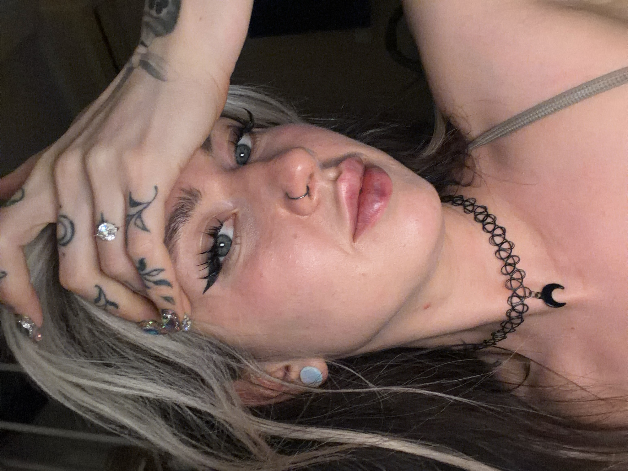 A close-up, tilted, high-angle shot of a woman. She has fair skin, blue eyes, and silver hair with dark roots. The woman is wearing makeup, including eyeliner, lipstick, and a nose ring. Her right hand is raised towards her forehead, revealing finger tattoos and a large diamond ring. She also has manicured nails with an iridescent design. The woman is wearing a tattoo choker with a crescent moon charm. A thin gray strap of an unseen garment is visible over her shoulder. The environment is dark and undefined. This image was not taken in Budva, Montenegro.