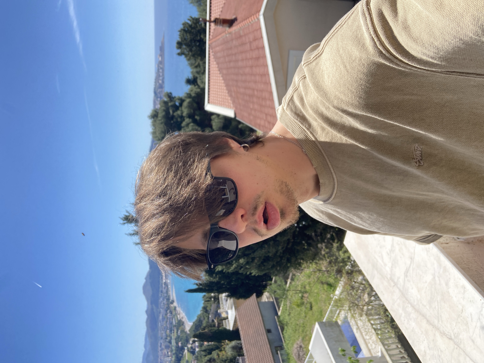 A young man wearing sunglasses and a tan t-shirt takes a selfie outdoors. He stands on a white balcony, overlooking a landscape of trees, buildings with red tile roofs, and a coastline. The sky is blue, and the sea stretches into the distance. Šušanj, Montenegro, is visible in the background along the coastline.