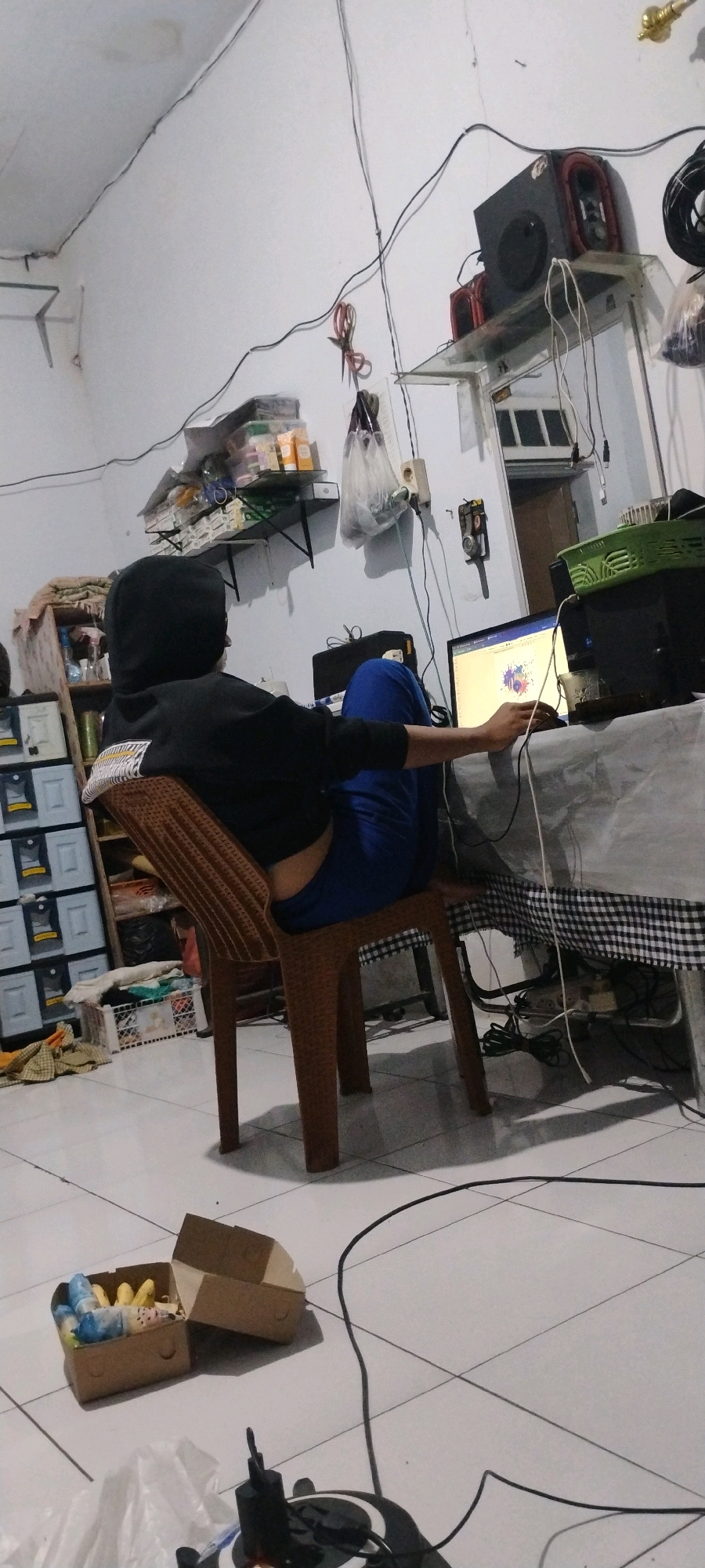 In an indoor scene, likely a residence in Mumbulsari, Indonesia, a person wearing a black hooded jacket and blue pants is seated on a brown plastic chair. The individual is facing away from the camera, towards a laptop computer that is open on a table covered with a gray and black checkered tablecloth. The laptop screen displays a colorful abstract image. To the left of the person, a set of white plastic drawers with yellow accents is visible. Further to the left, a brown shelf unit holds various items, including bottles and what appear to be hygiene products. Above this shelf, a smaller shelf is attached to the white wall, also holding several bottles. A pair of scissors is hanging from a string on the wall above the smaller shelf. To the right of the laptop, a black speaker is mounted on a clear shelf. Various cables are draped across the wall and floor, suggesting electronic equipment is in use. In the foreground, on the tiled floor, a cardboard box is open, containing what appear to be small stuffed toys. Another empty cardboard box lies next to it. The walls are painted white, with some visible discoloration or marks. The lighting appears to be artificial, creating shadows on the floor. The overall impression is a cluttered, lived-in space.