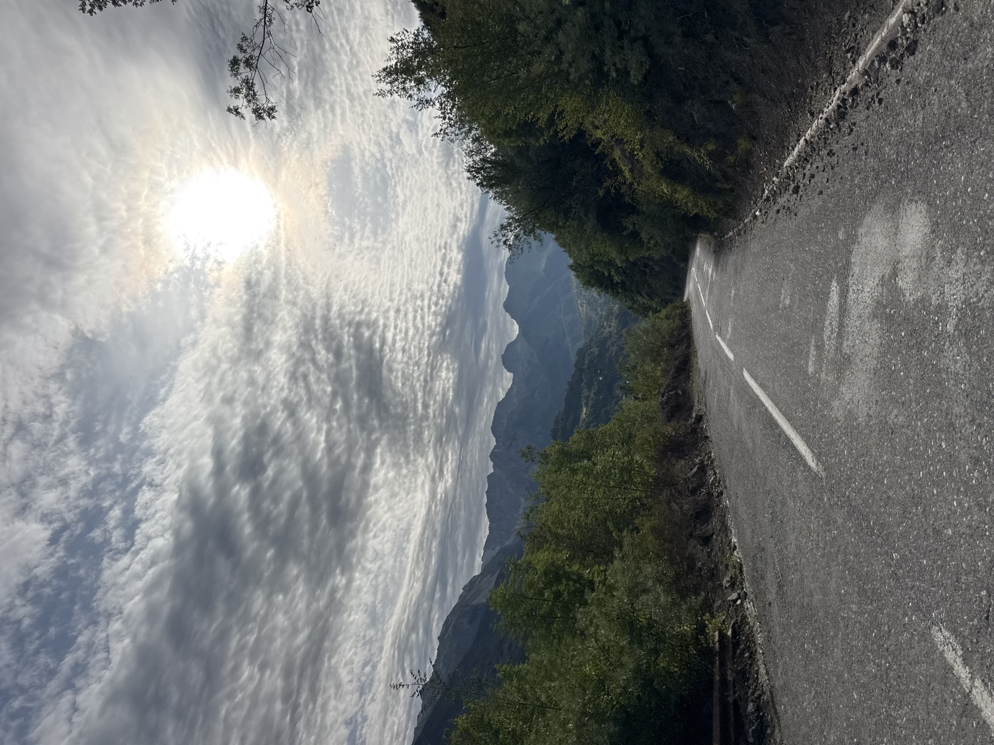 An asphalt road winds through a mountainous landscape under a cloudy sky. The sun shines brightly through a break in the clouds, creating a halo effect. Lush green trees line the sides of the road, and in the distance, a range of dark, jagged mountains rises against the horizon. The road has a white line marking the center and is slightly wet in places. Debris is visible along the roadside in some areas. The scene appears to be in Dragovića Polje, Montenegro.