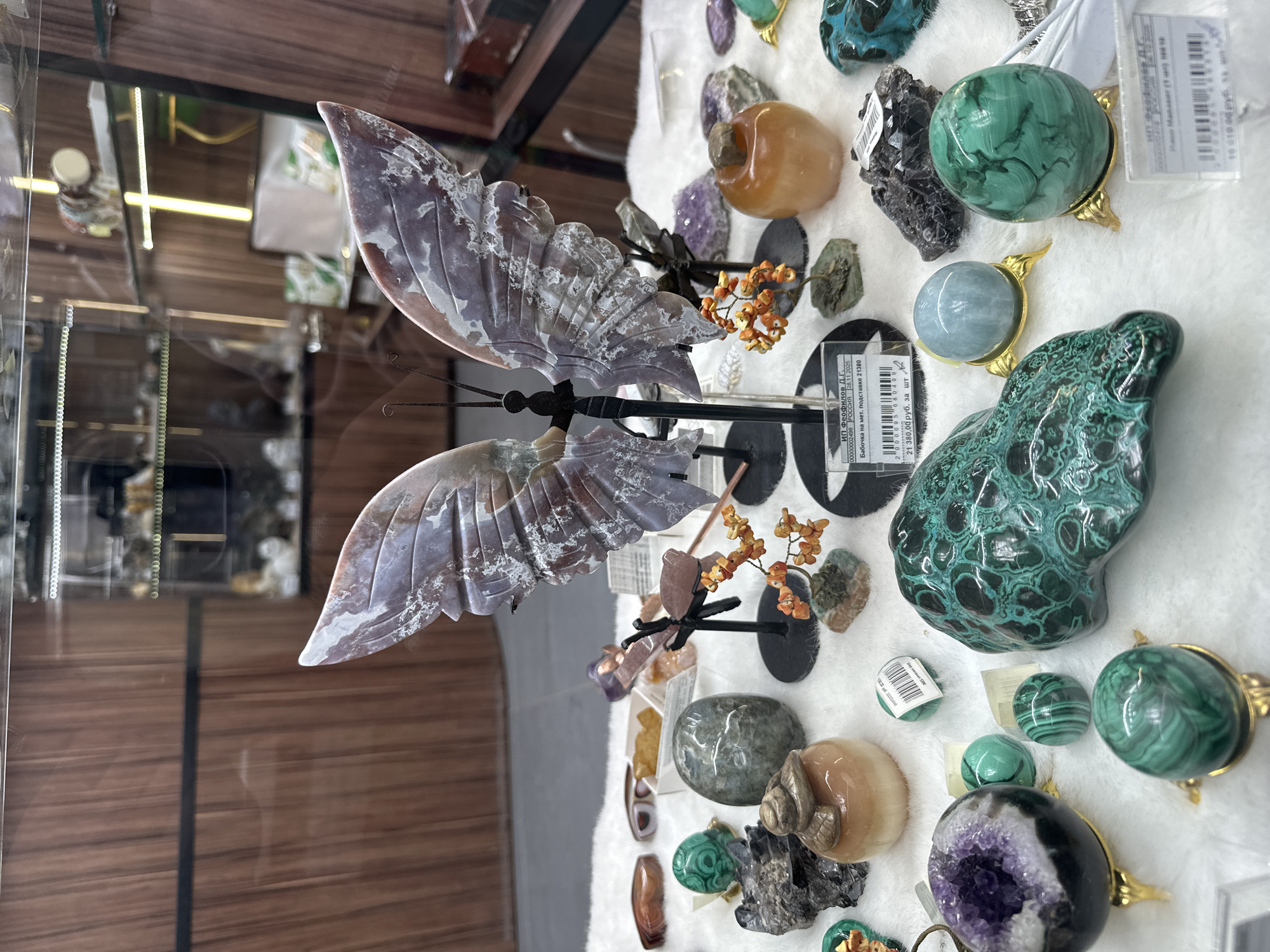 This image captures a retail display of various mineral and crystal items, likely in a shop in Bolshoye Savino, Russia. The central and most prominent subject is a large, intricately carved butterfly made from a patterned stone, possibly jasper or agate, with wings displaying shades of grey, purple, and brown with white inclusions. It is mounted on a black metal stand.
Around the butterfly, a diverse collection of polished and raw minerals is arranged on a soft, white display surface. Notable items include multiple pieces of green malachite, presented as spheres (some in golden stands) and a larger, irregularly shaped sculpture. There is also a light blue/grey sphere in a golden stand, an amethyst geode displaying purple crystals, and various other tumbled stones in hues of orange, brown, and clear. Small decorative clusters resembling orange berries on black stems are interspersed among the minerals.
Reflections on the left side of the image suggest the display is enclosed within a glass case, showing blurred views of wooden paneling and interior lighting fixtures of the shop.
Visible text on the price tags and labels provides specific information. One label identifies the vendor as "ИП Феофилов Д.Г." and the item as "Бабочка на мет. подставке" (Butterfly on a metal stand), with a price of "21 380,00 руб. за шт" (21,380.00 rubles per piece). Another price, "16 618.00 руб", and the country "РООССИЯ" (RUSSIA) with a date "28.11.2025" are also legible. The image depicts a static scene of goods for sale in an indoor retail setting.