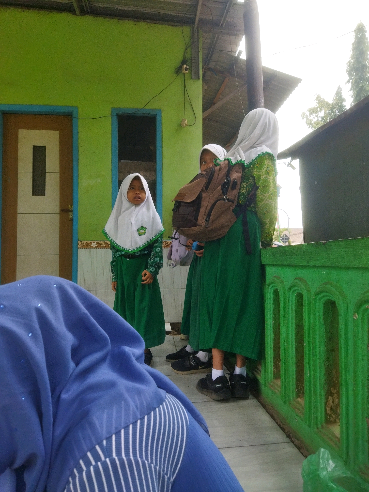 The image depicts an outdoor scene in Pasrepan, Indonesia, with four individuals visible. In the foreground, a person wearing a blue headscarf is partially visible. Behind them, three young girls, appearing to be students, are standing. The two girls on the right are wearing green school uniforms and white hijabs, with one carrying a brown backpack. The girl on the left is also in a green uniform and white hijab, and she is looking towards the viewer. The environment includes a lime green building with blue-trimmed windows and doors, a wooden roof structure, and a vibrant green railing. The ground is paved with light-colored tiles. The overall scene suggests a school setting or a gathering outside a school.