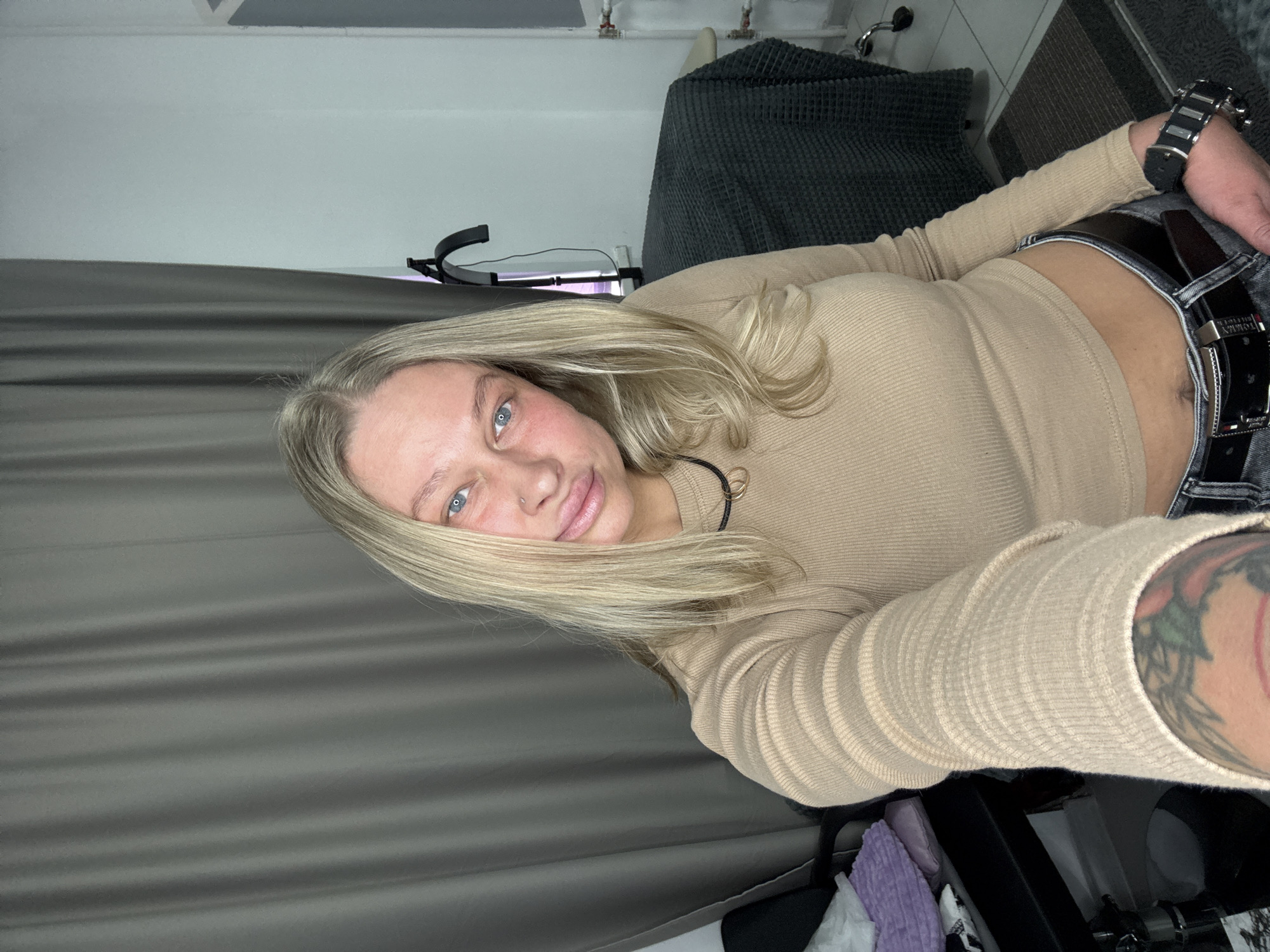 A young adult woman with long blonde hair and blue eyes takes a selfie in what appears to be an indoor setting, possibly a room in Perm, Russia. She looks directly at the camera with a gentle, slightly smiling expression, featuring a nose piercing and full lips. She is wearing a light beige ribbed long-sleeved crop top that reveals a sliver of her midriff, dark jeans, and a black belt. The belt buckle clearly displays the text "TOMMY HILFIGER". On her left wrist, she wears a large, chunky watch with a black strap and silver-toned casing. A colorful tattoo, featuring what looks like green leaves and possibly red fruit or flowers, is visible on her right arm.
The background includes a dark gray curtain on the left side and a dark gray textured blanket or cover on a piece of furniture in the upper right. White pipes are visible on a wall in the upper right, and a black light stand or tripod is positioned behind her. Various indistinct objects, including some purple and white fabric, are partially visible in the lower right corner, along with a portion of a dark mat or rug. The lighting suggests an interior setting, possibly enhanced by artificial light.
