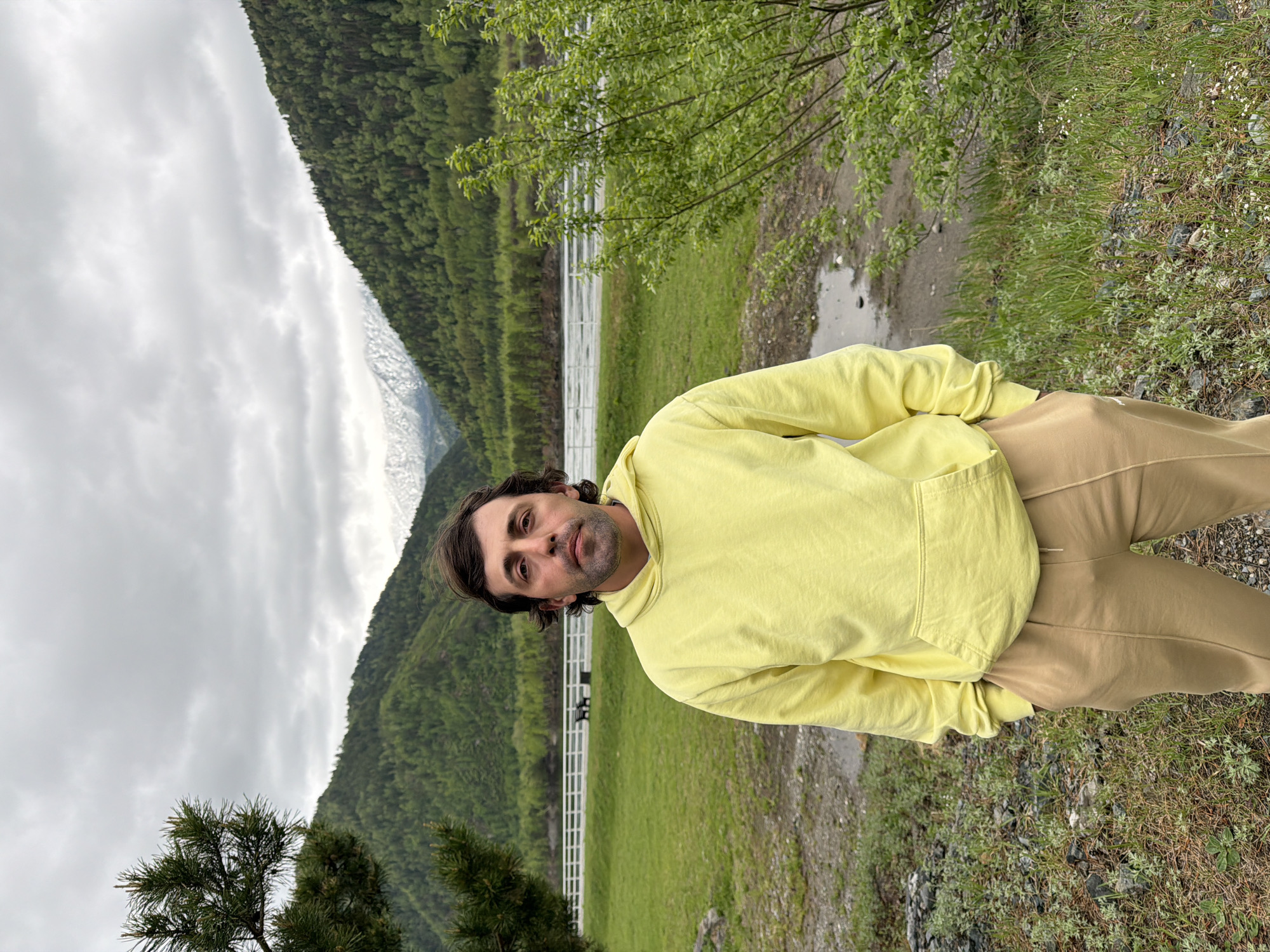 A man is posing for a photo outdoors. He is wearing a yellow hooded sweatshirt and light brown pants. Behind him, a forested mountain rises under a cloudy sky. A white fence runs along the grassy area. The location is not identifiable within the city of Cheremushki, Russia, based solely on the visual information provided.