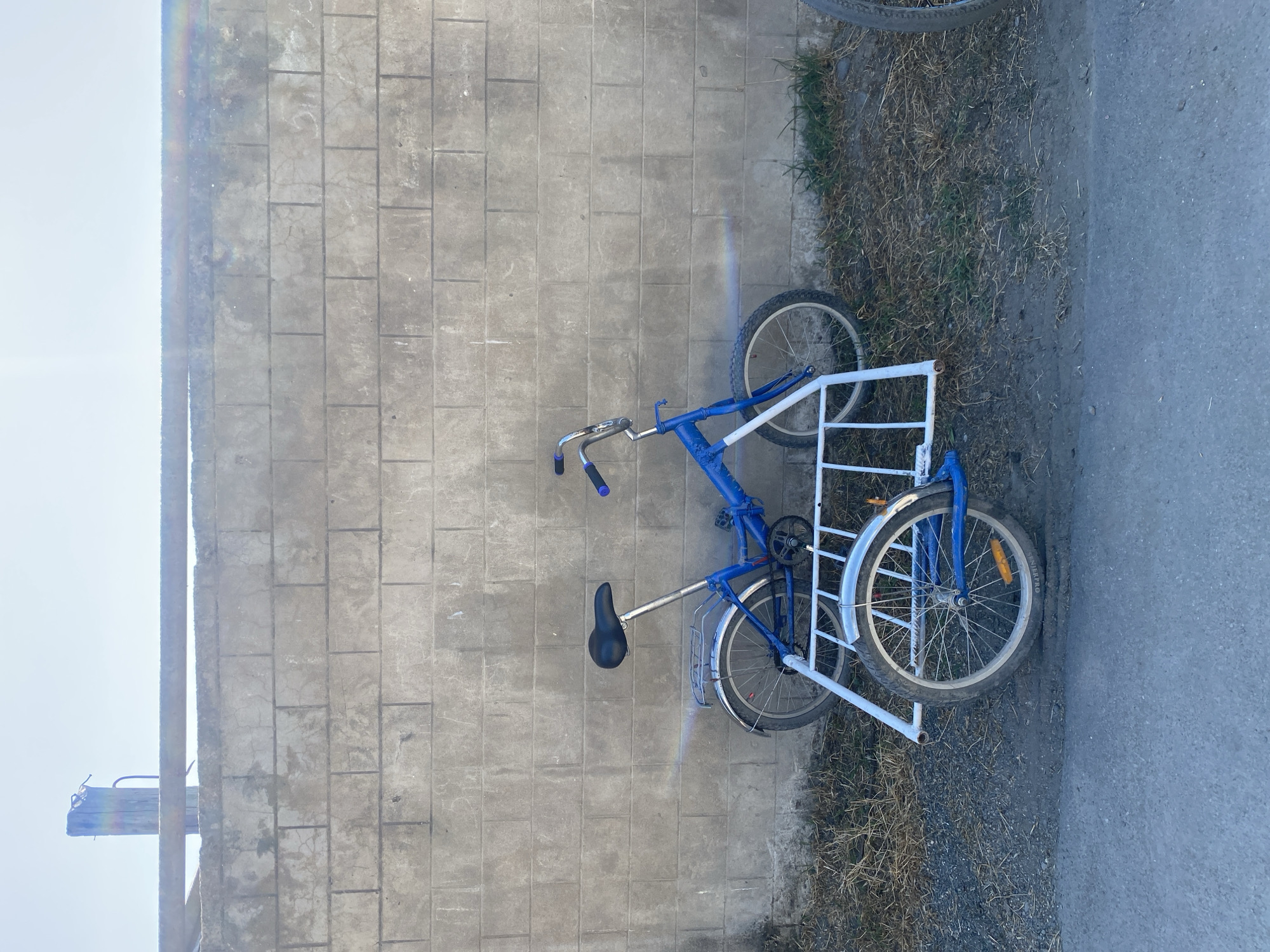 A blue tricycle with a white frame is parked against a cinder block wall in an outdoor setting. The tricycle has a seat, handlebars, pedals, and wheels, with a cargo area constructed from white tubing at the rear. The wall is made of large, rectangular cinder blocks and shows signs of wear. The ground in front of the wall is a mix of gravel and dry grass. The sky above is clear and bright. The scene appears to be located in a rural or semi-urban area.