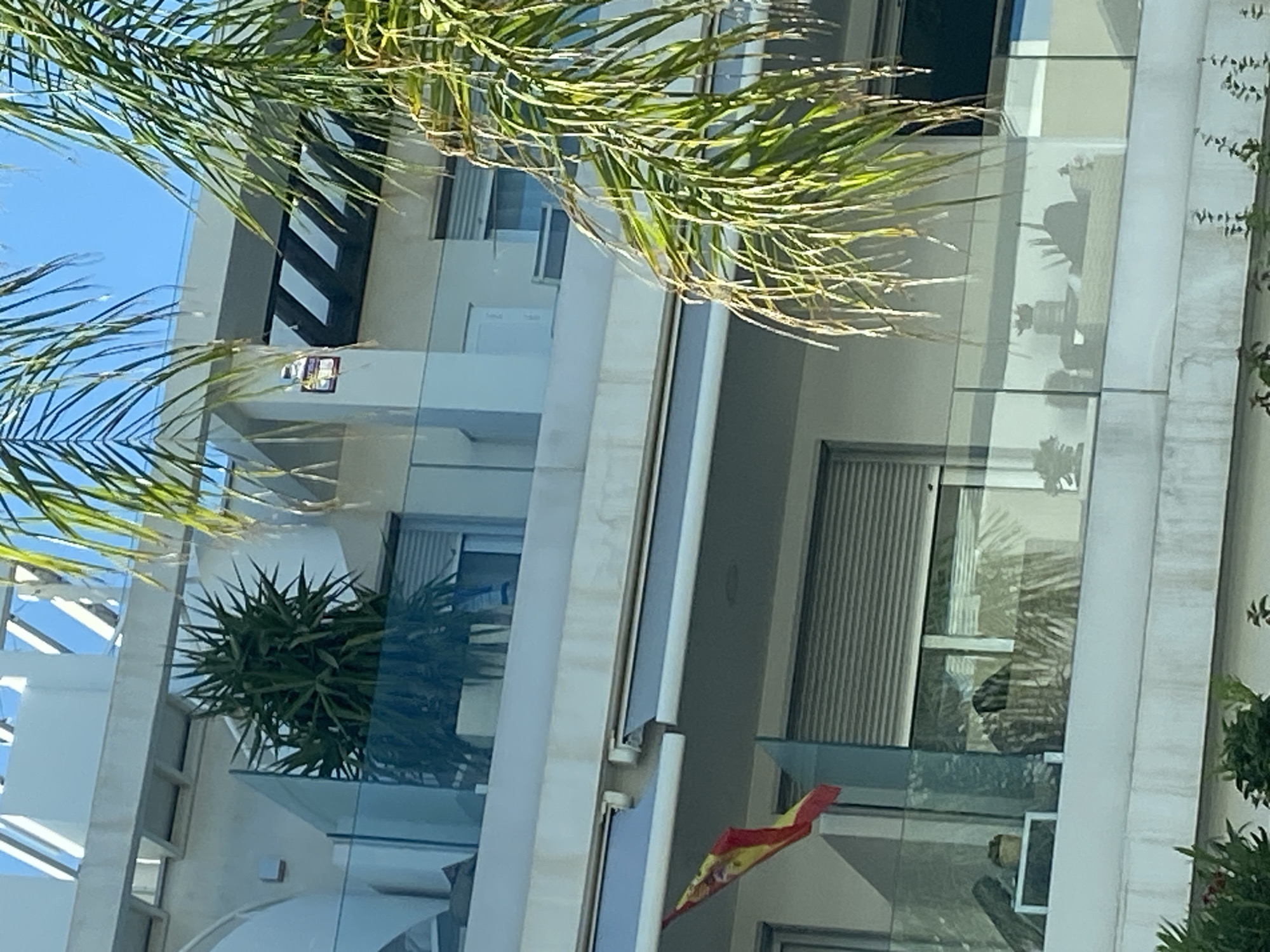 This is a close-up, eye-level, vertical, exterior shot of a modern building with multiple balconies, likely a residential apartment building, located in the city of La Antigua, Spain. The building features white concrete walls, glass railings on the balconies, and roll-down shutters on some windows. A Spanish flag is visible on the left side of a balcony, attached to a pole. Plants, including a large spiky plant and palm fronds, are prominently featured in the foreground and partially obscuring the view of the building. The sky is visible and appears clear and blue. There are no people clearly visible in the image. The overall impression is that of a bright, sunny day in a coastal or Mediterranean location.