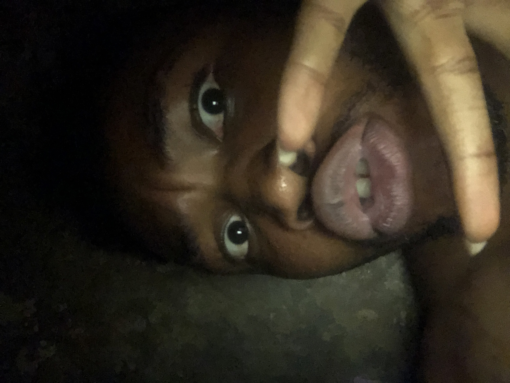A close-up, low-light image depicts a person with dark skin, possibly a male, partially facing the camera. The person's eyes are wide, and their mouth is open, with the tongue visible. A hand with long fingers is positioned near the person's face. The background is dark and indistinct, making it impossible to discern any details about the location. The image does not offer any clues about the city.