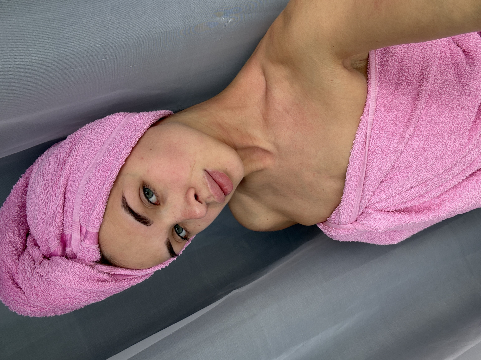 A woman with a pink towel wrapped around her head and shoulders lies on a gray surface. The woman has fair skin, green eyes, and dark eyebrows. Her head is tilted to the side, and she is looking directly at the camera. The background is out of focus. The location in Sarapul, Russia is not discernible from the image.