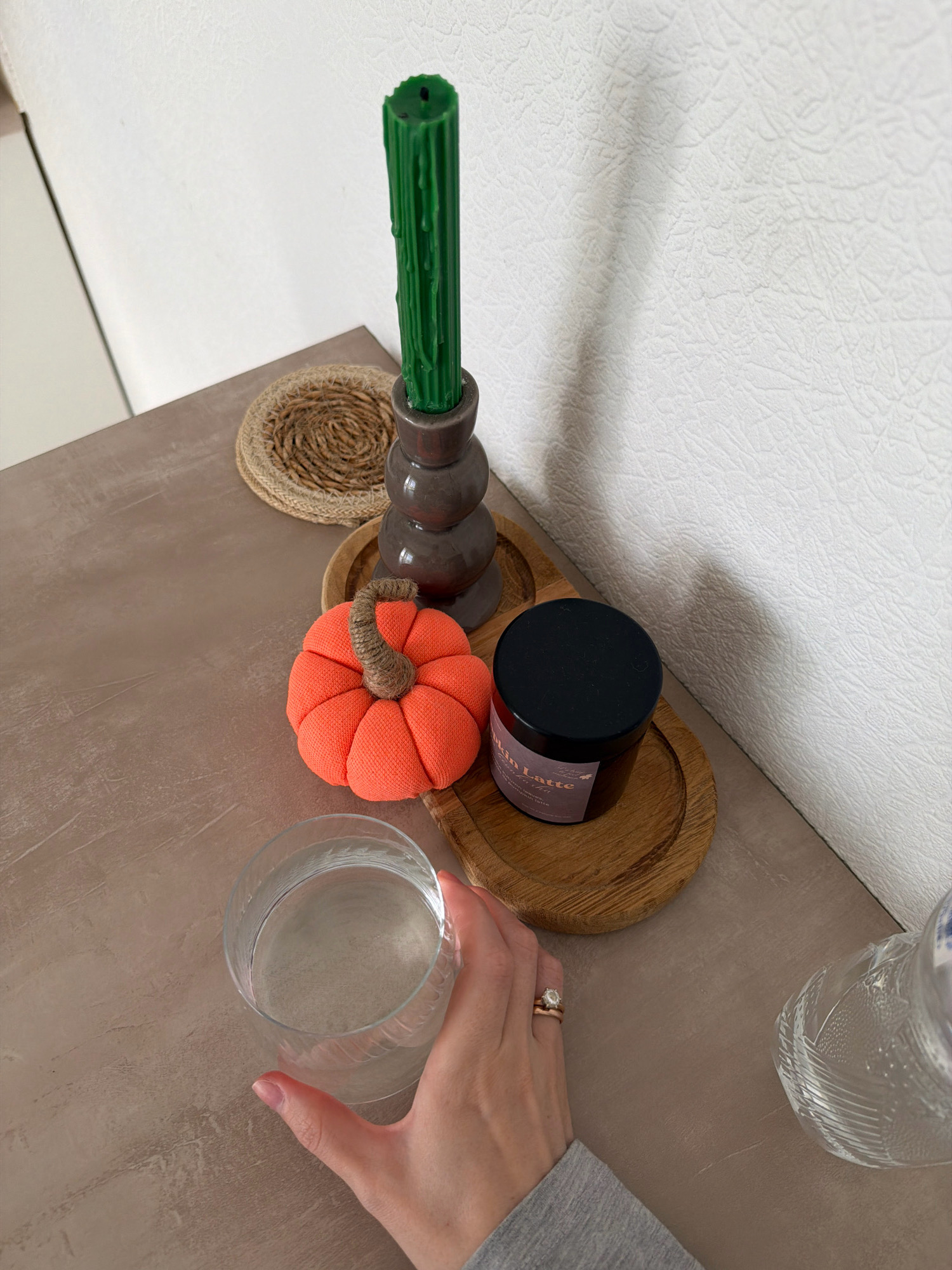 The image shows a close-up, high-angle view of a table surface with several decorative items. A person's hand, adorned with two rings, is holding a clear glass filled with a light-colored liquid. To the left of the glass is an orange fabric pumpkin with a textured brown stem. Behind the pumpkin is a dark gray, multi-tiered candle holder, topped with a tall, ridged green candle. A round, woven placemat sits behind the candle holder. To the right of the pumpkin and candle holder is a small, dark jar with a black lid, resting on a round, wooden coaster. The jar has a label that reads "Pumpkin Latte" and other text in a smaller font. To the far right, part of a clear, textured glass bottle is visible. The table surface appears to be a neutral, textured material. The background consists of a textured white wall. The scene is an indoor still life. No location in Kyiv, Ukraine is specified or inferable.