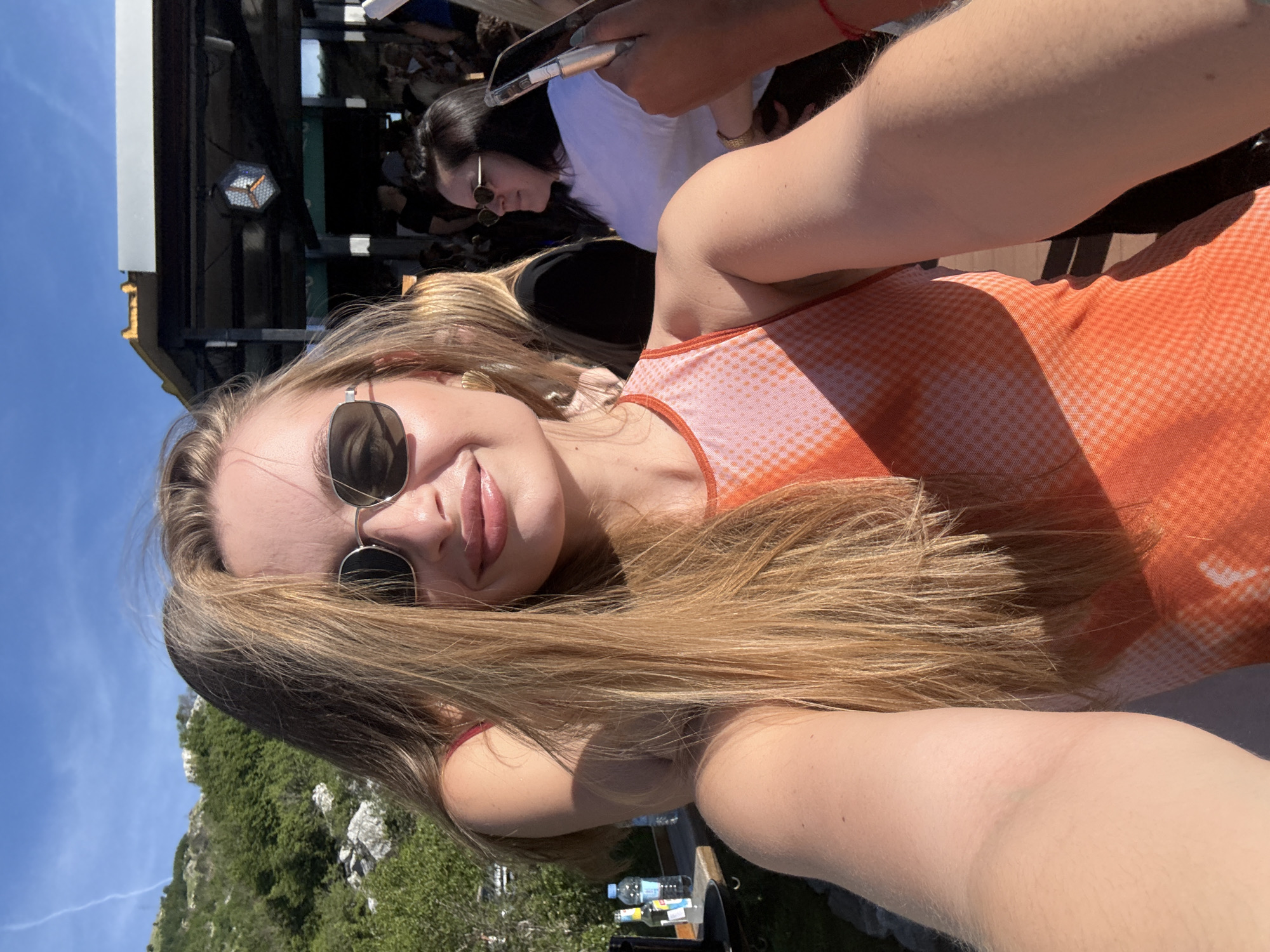 A young woman with long blonde hair, wearing sunglasses and an orange and white patterned top, takes a selfie outdoors. She is smiling and has glossy lips. Behind her, a blurred background shows a mountainous landscape with green trees under a clear blue sky, a shaded outdoor seating area with people, and a table with bottles of water. The woman's arm is visible in the foreground, resting on her shoulder. The scene suggests a casual social gathering in a scenic location.