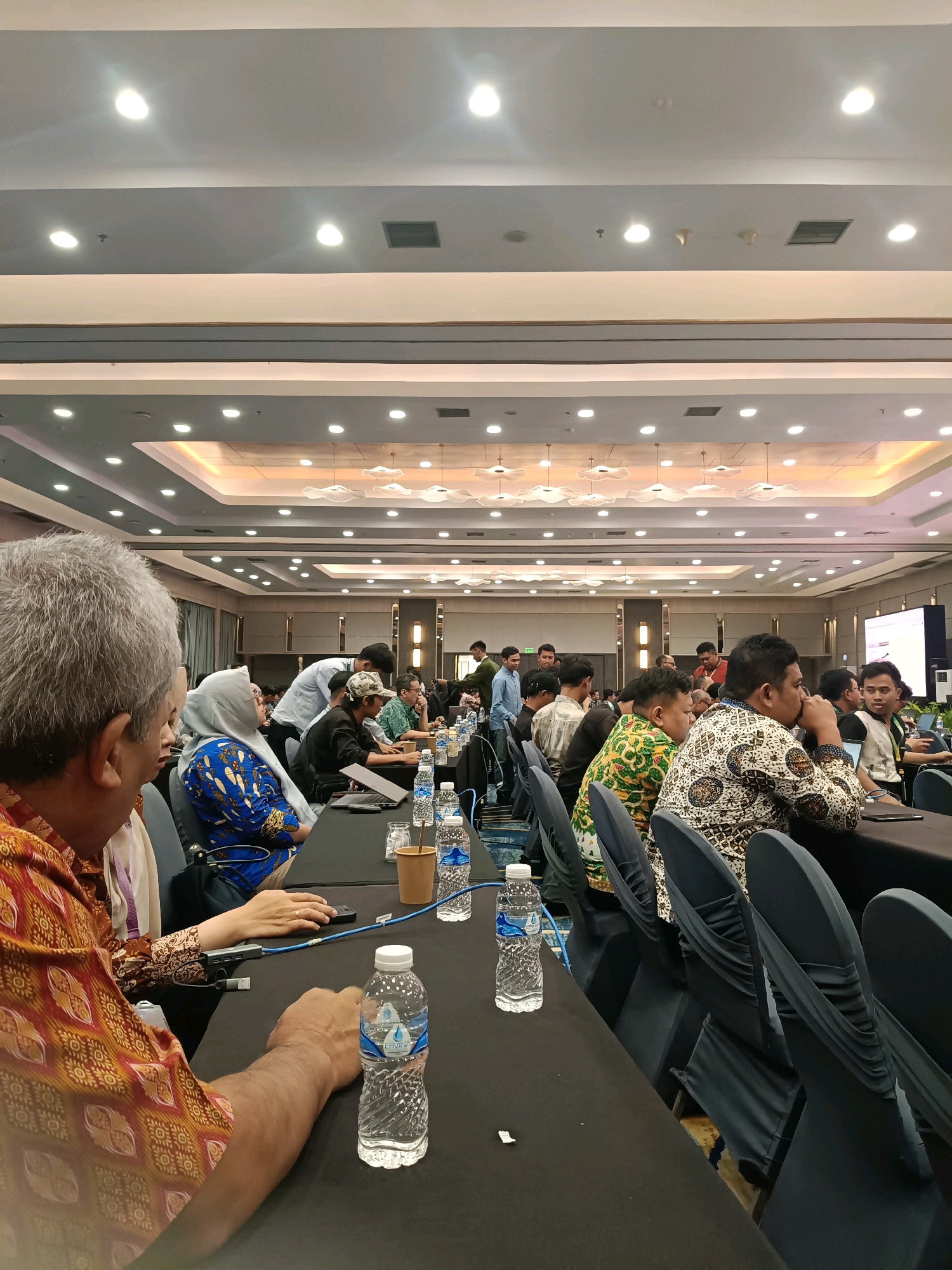 The image shows a conference or seminar event at the JIExpo Kemayoran in North Jakarta, Indonesia. Attendees are seated at long tables covered with black tablecloths, facing a stage or presentation area that is not fully visible. The room has a high ceiling with multiple tiers of lighting fixtures, including recessed spotlights and decorative ceiling elements. On the tables, there are several bottles of water, laptops, and personal belongings. Some attendees are interacting with their devices, while others are engaged in conversation or listening to a speaker. The overall atmosphere suggests a professional or academic gathering.