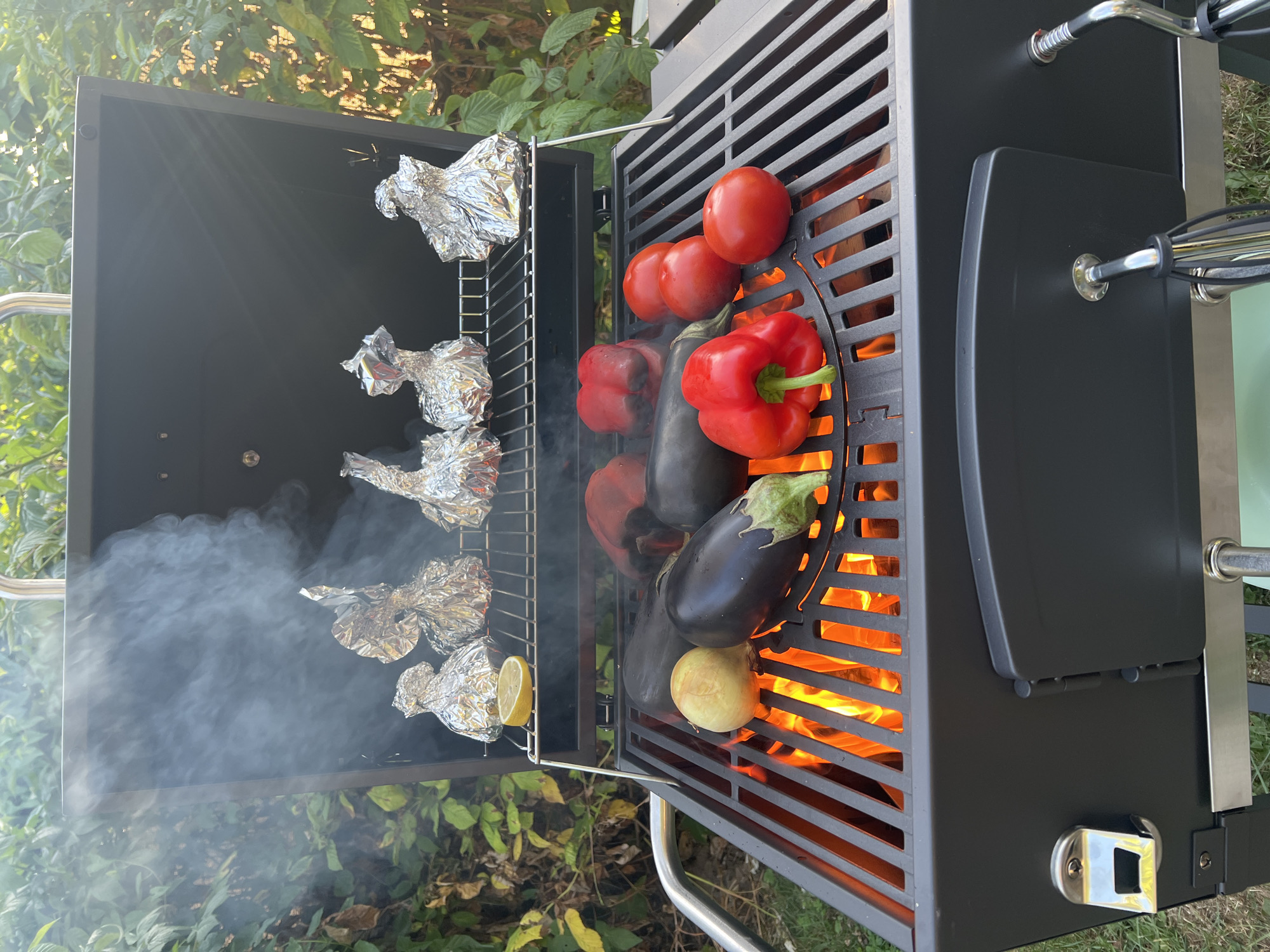 This image depicts an outdoor barbecue in the city of Denderleeuw, Belgium. A black charcoal grill is actively in use, with visible flames and smoke rising from the grates. On the lower grate, several vegetables are being grilled: two eggplants, one whole onion, two red bell peppers, and three tomatoes. Above, on a raised rack, are five foil-wrapped parcels, suggesting food being cooked indirectly. A single lemon slice is also placed on this upper rack. The background shows a lush green garden with dense foliage, indicating a residential or private outdoor space. The lighting suggests it is daytime, possibly late afternoon, with sunlight filtering through the leaves. There are no people visible in the frame.