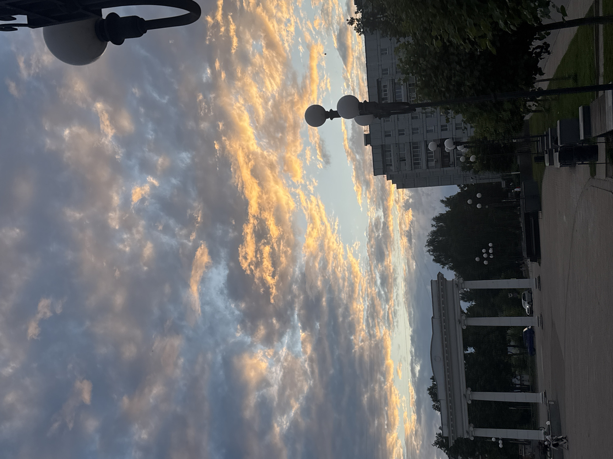 The image depicts a dusk scene in Chaikovsky, Russia, likely in a public square or park. A large, ornate archway with classical columns stands prominently on the left side of the frame, casting shadows on the paved ground. Behind the archway and to the right, a multi-story residential building is visible, its windows reflecting the fading light. Several lampposts, some with multiple spherical lights, line a pathway in the foreground, suggesting an urban setting. Trees, some appearing to be deciduous and in full foliage, form a backdrop and border the scene. The sky is dramatic, filled with scattered clouds illuminated by the setting sun, creating streaks of orange and yellow against a blue and grey canvas. A few cars are present on a road visible through the archway. A couple of individuals are seen walking in the distance on the left. The overall atmosphere is serene and twilight-like.