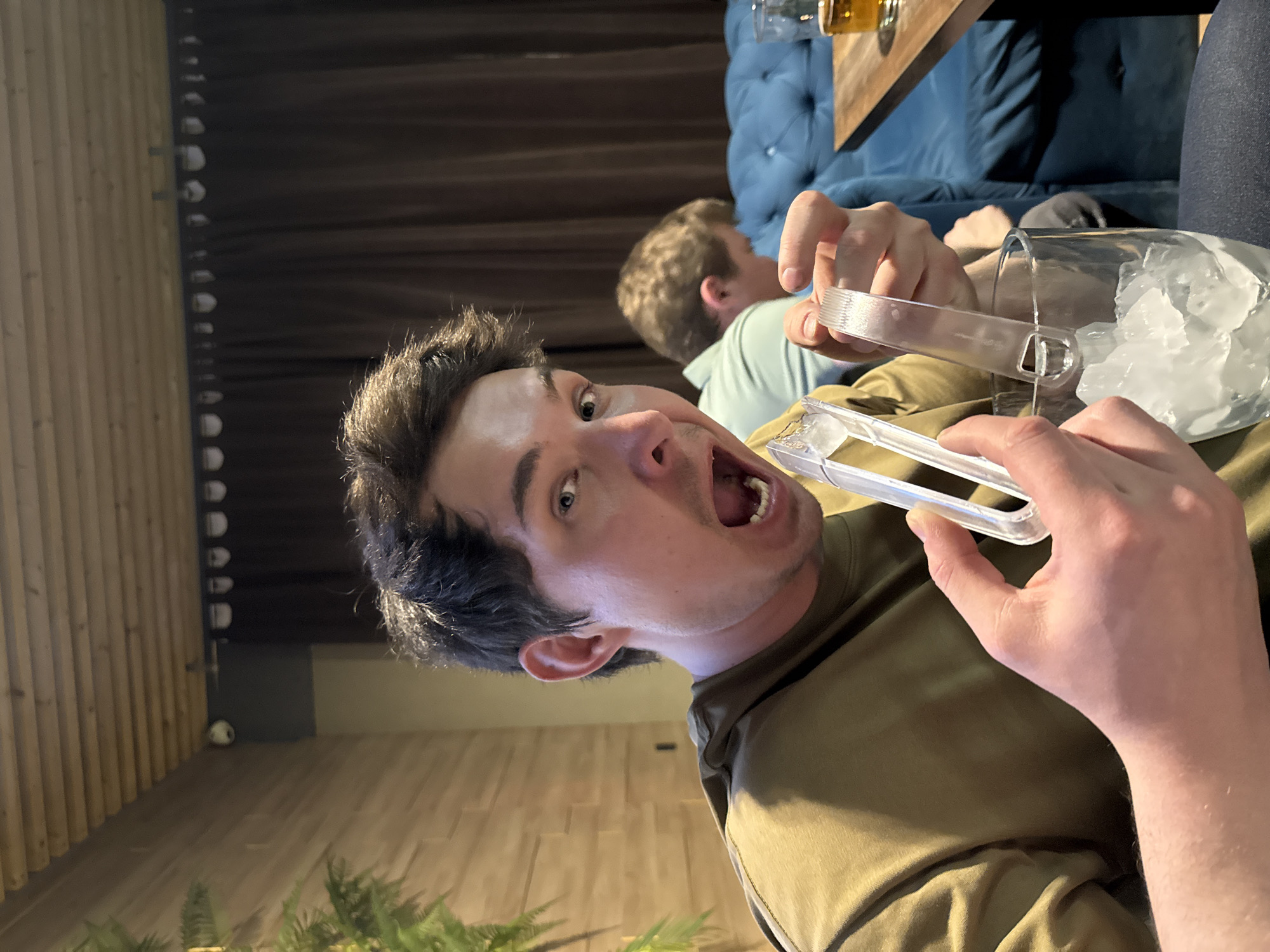 A man with dark hair and an open mouth holds an ice tong near a glass filled with ice cubes, seemingly at a bar or lounge. Another person sits in the background, slightly out of focus. A blue tufted sofa is also visible. The image was reportedly taken in the city of Chaikovsky, Russia, but this cannot be confirmed from the image alone.