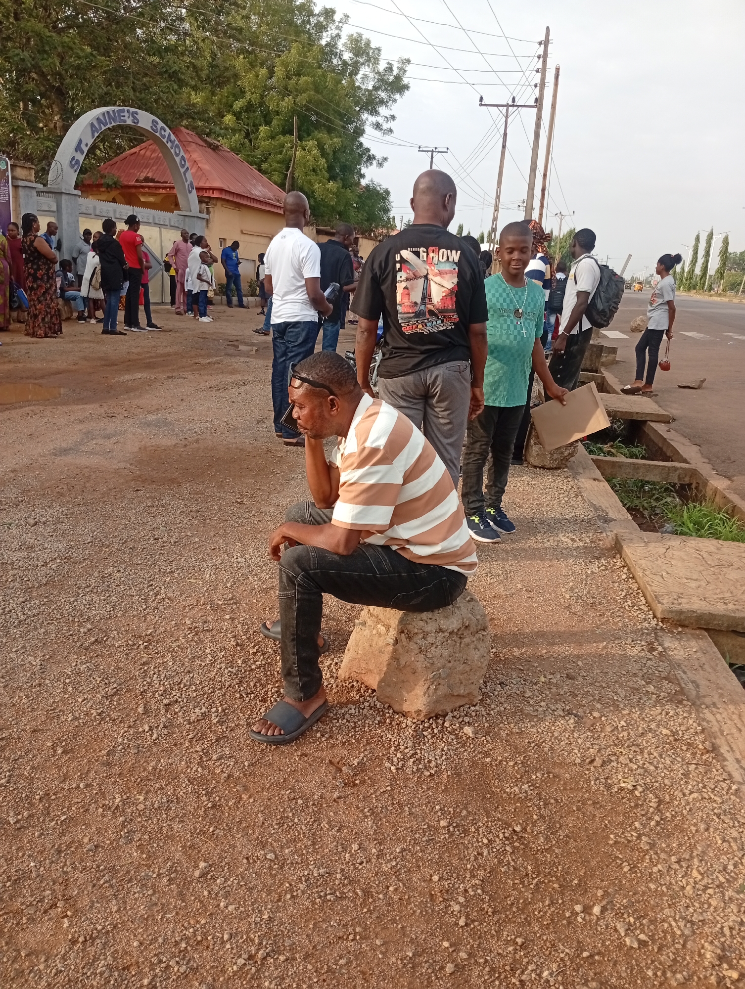 In Barden Yana, Nigeria, a man sits on a stone near a road, appearing contemplative. He wears a striped shirt, dark pants, and sandals. Behind him, a group of people stand near "ST. ANNE'S SCHOOL" entrance. Some are looking towards the school while others are walking along the road, several buildings and utility poles are visible in the background.